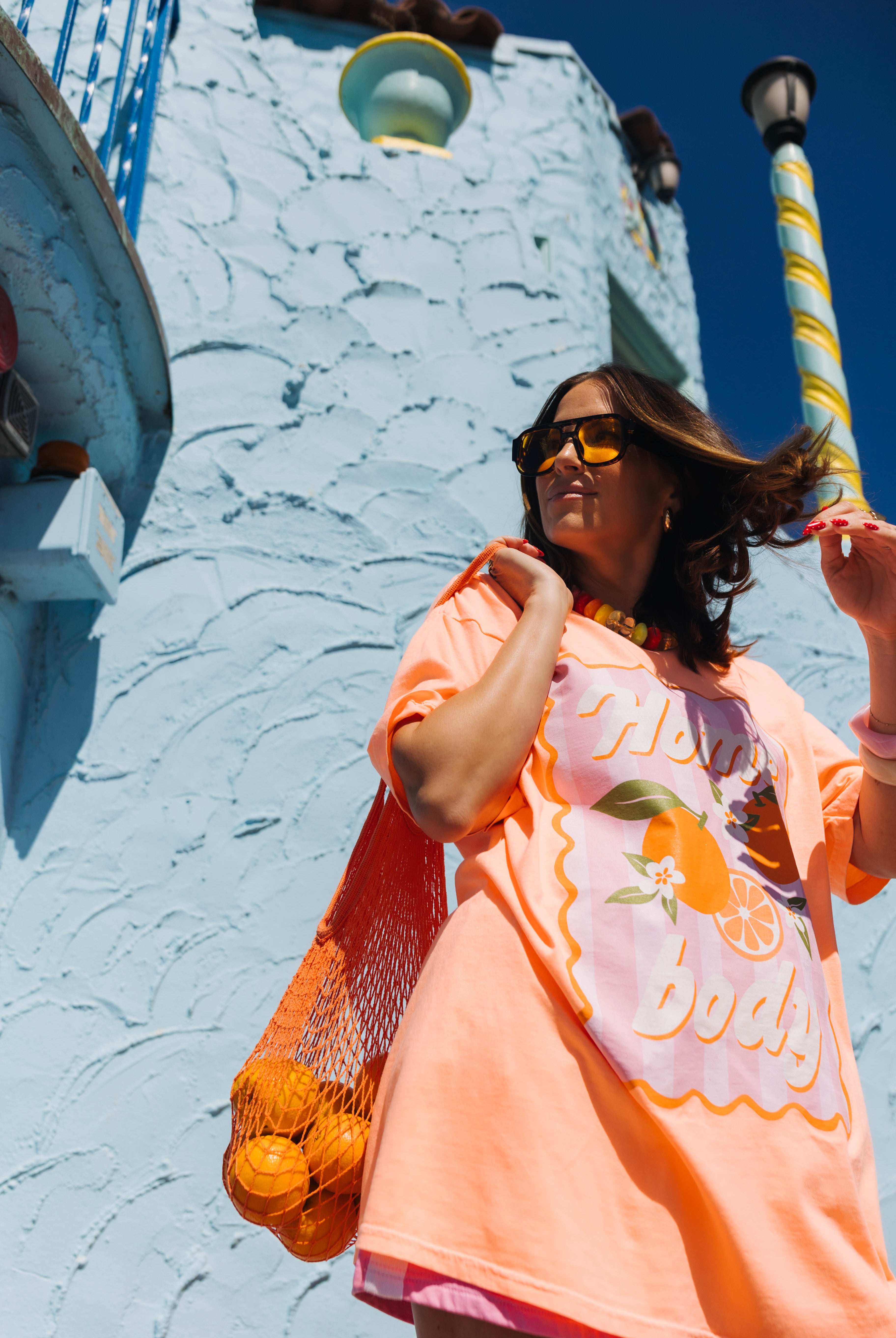 A woman wearing a bright orange graphic t-shirt and yellow-tinted sunglasses, carrying an orange mesh bag of oranges against a light blue textured stucco wall.
