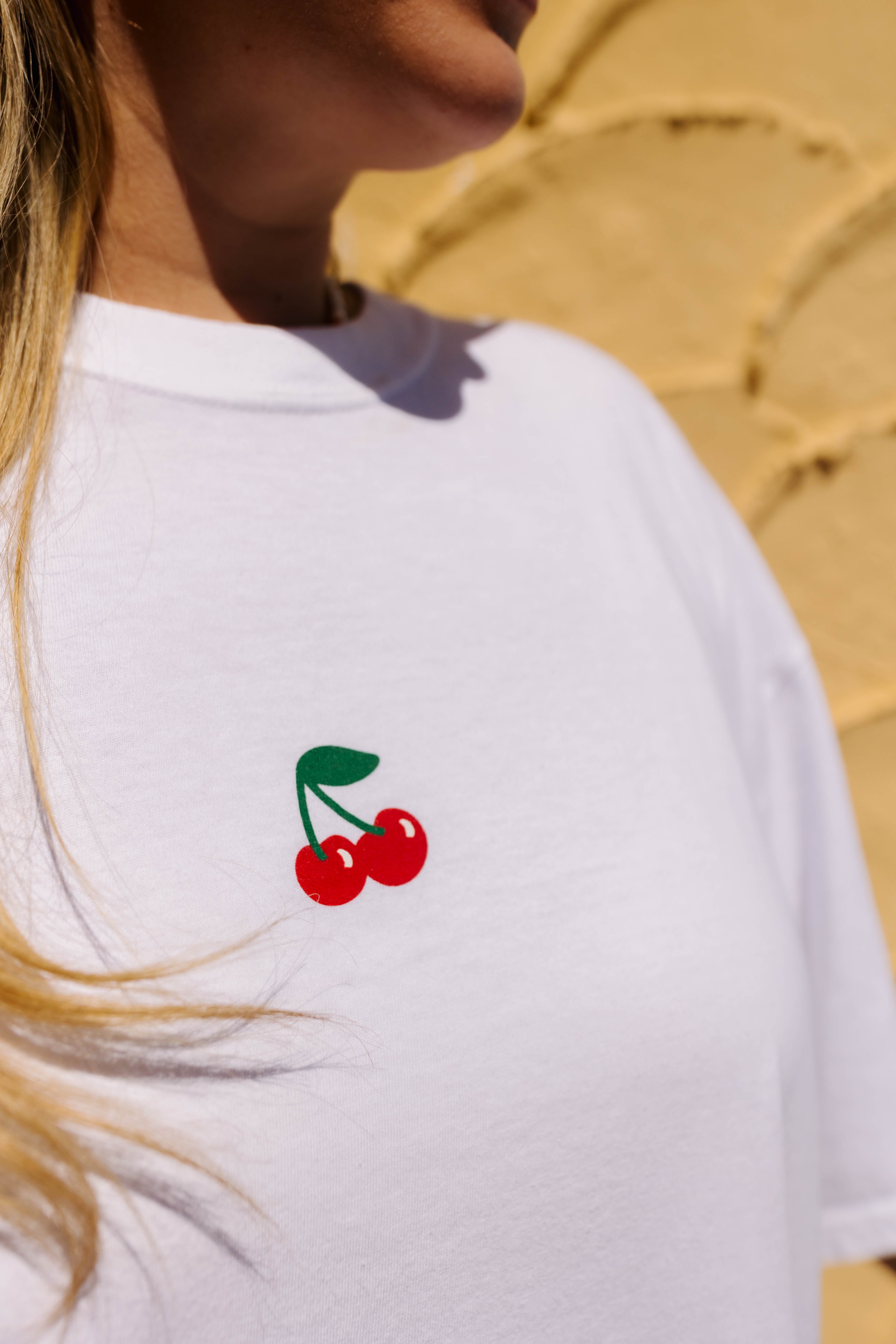 A close-up of a person wearing a white short-sleeve t-shirt with a small red cherry and green stem graphic on the left chest, set against a sandy beige background.