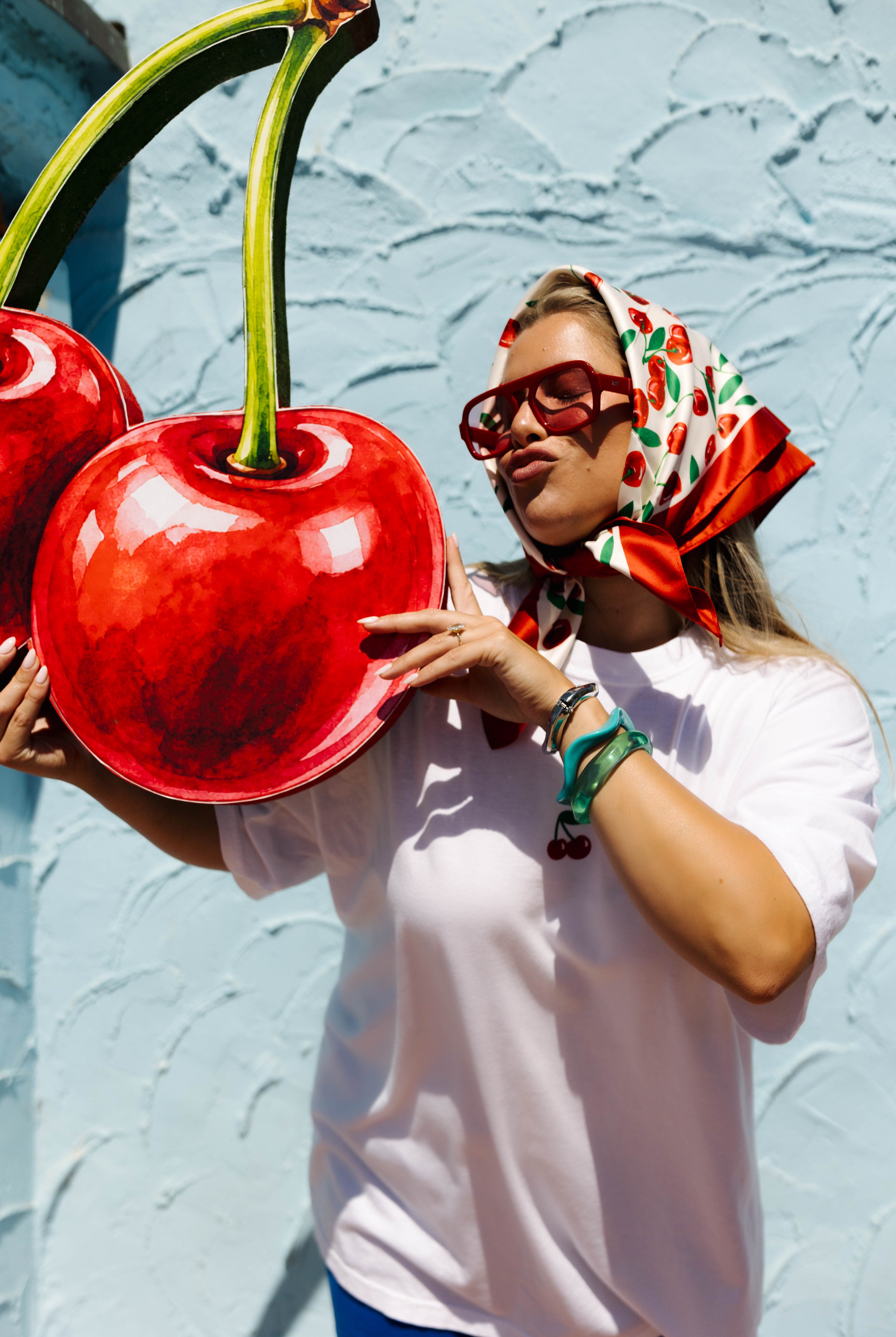 A woman wearing a white t-shirt, a red and green cherry-patterned headscarf, and red sunglasses holds a giant red cherry sculpture in front of a light blue textured wall.