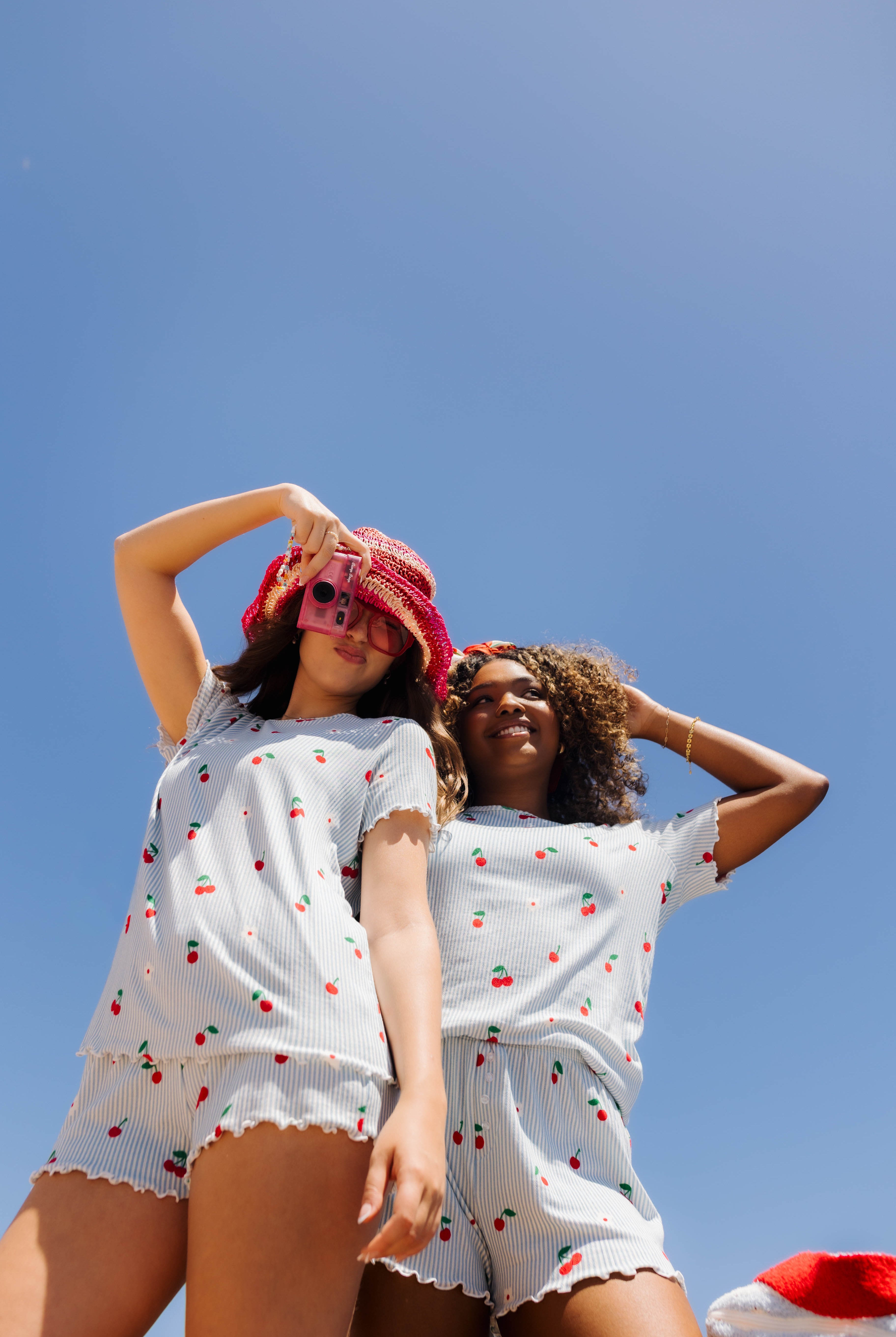 Two women wearing matching white short-sleeve pajama sets with a red and green cherry print, posing against a clear blue sky.
