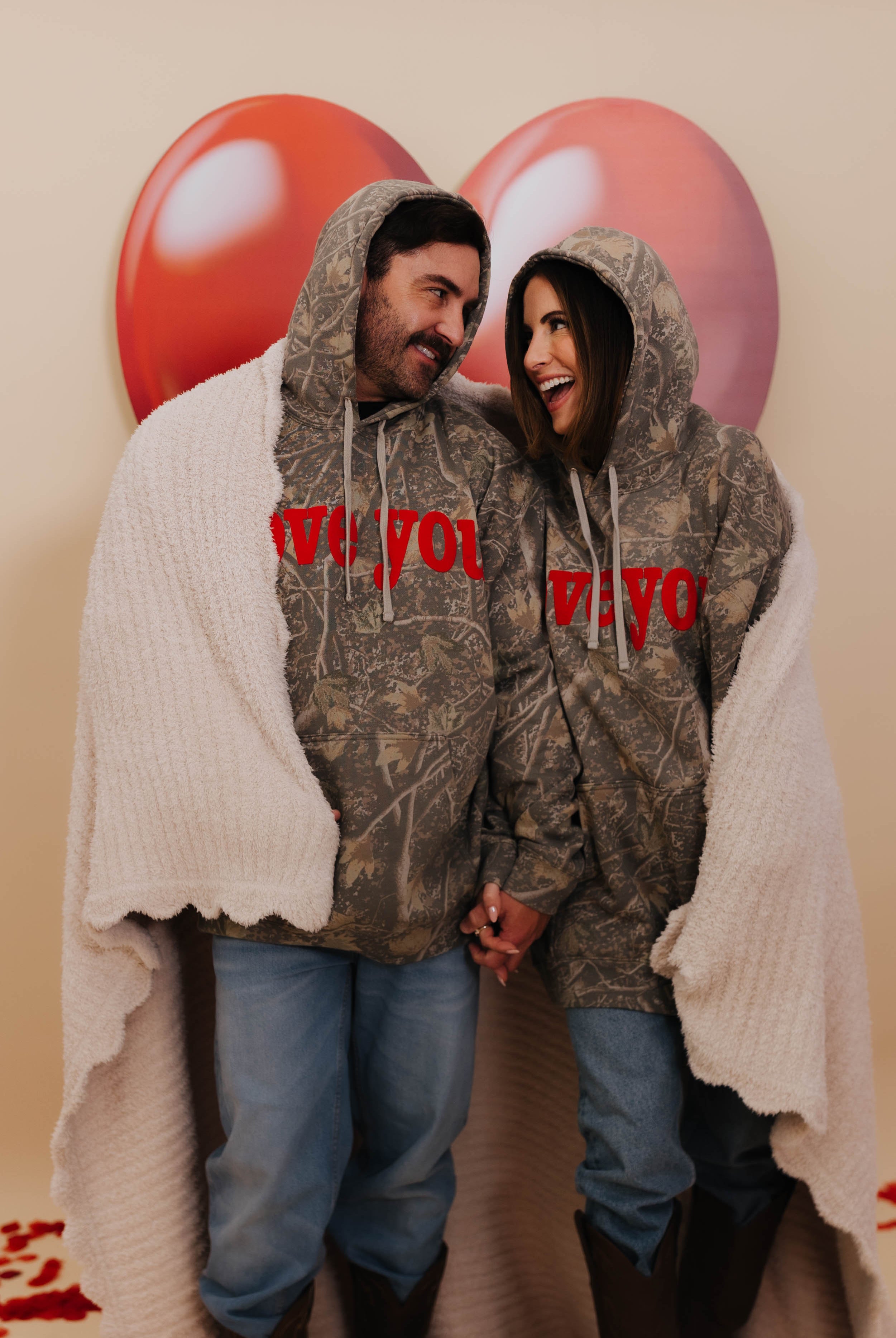 Two people wearing matching camouflage hoodies with 'love you' text, standing against a beige wall with heart-shaped balloons and wrapped in the scalloped blanket.