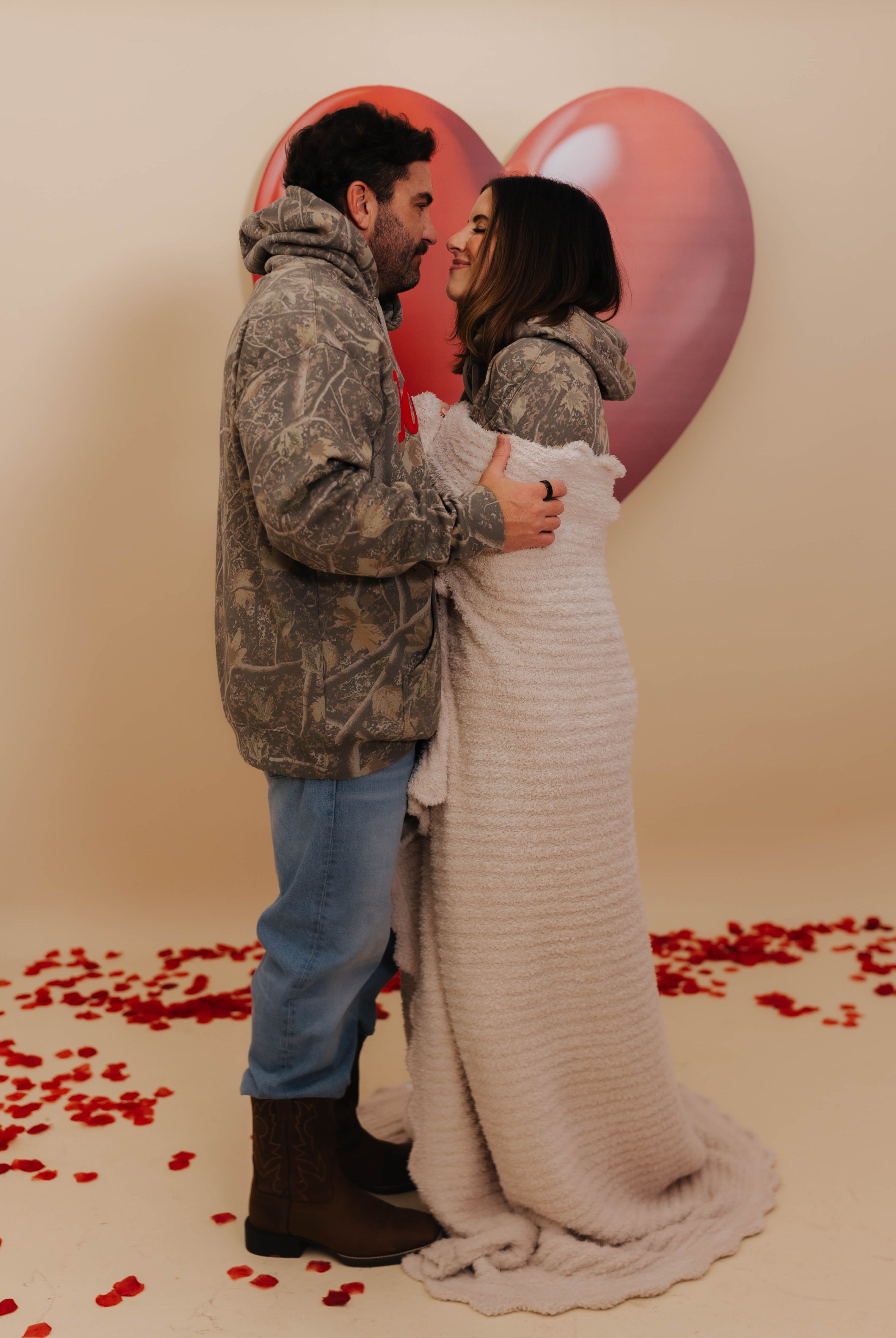 Couple embracing in front of heart-shaped balloons with red rose petals on a beige background with Krista wrapped in the blanket