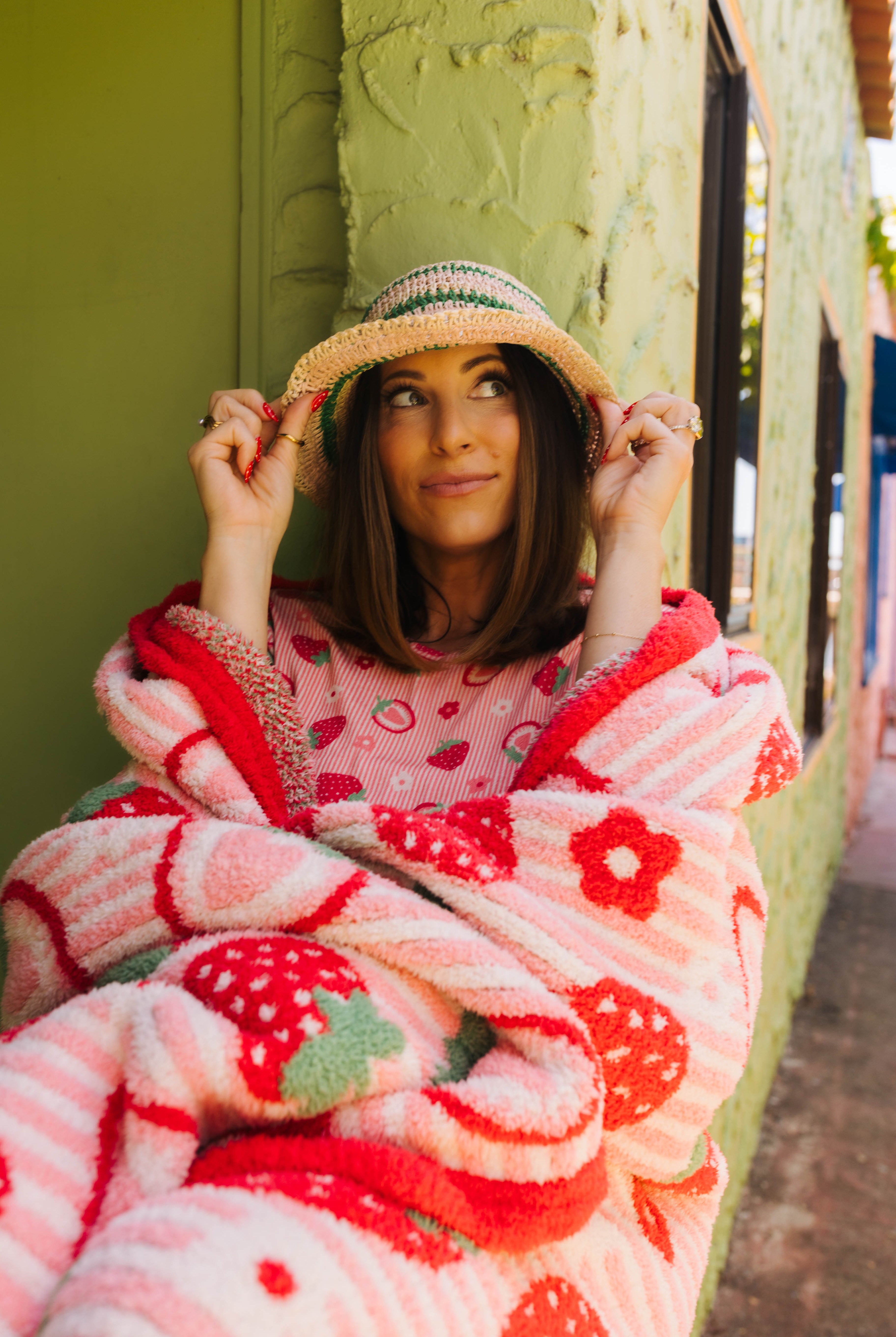 A woman wrapped in a pink and red strawberry-patterned plush blanket, wearing a straw hat, leaning against a textured olive green wall.