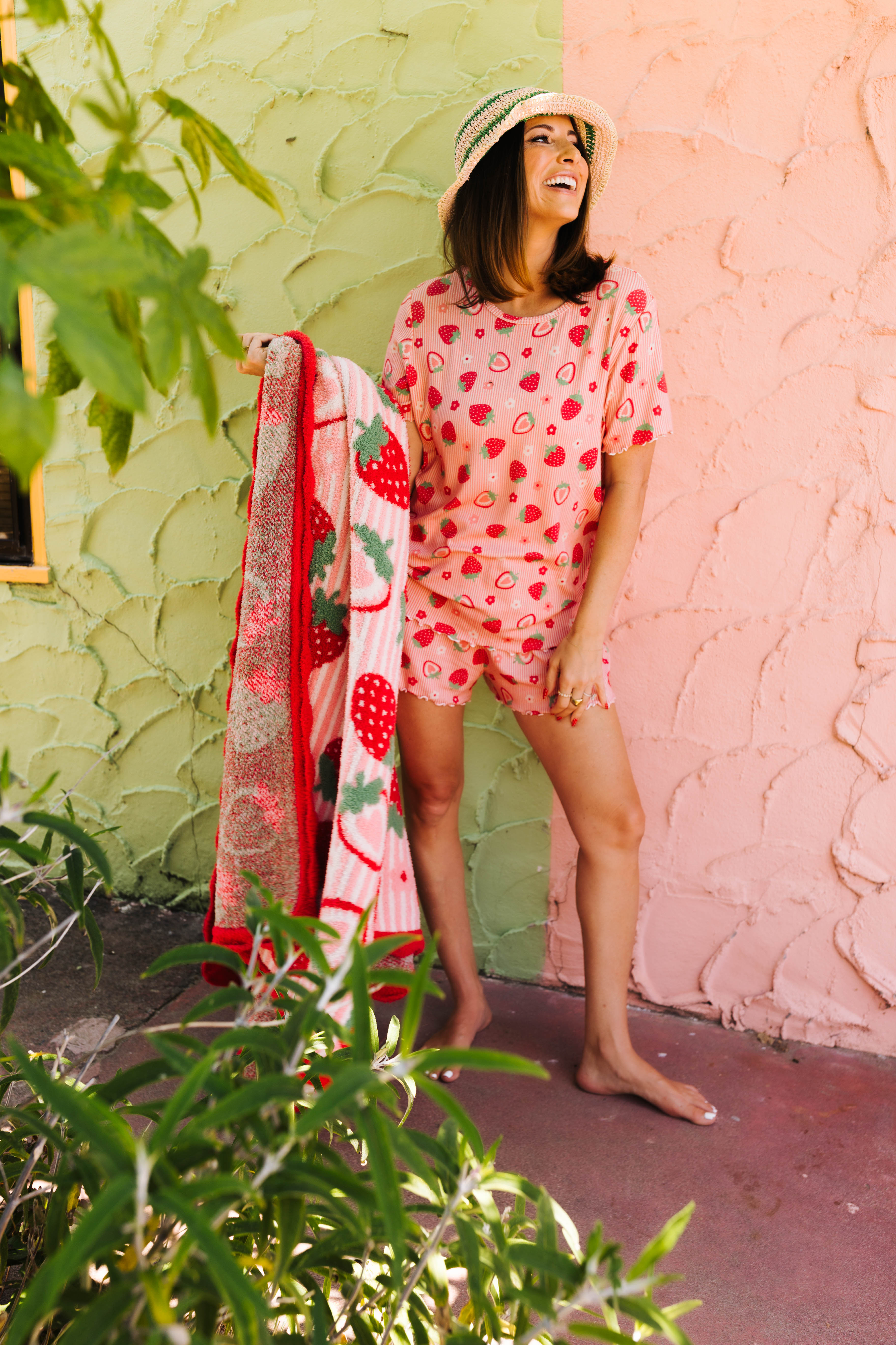 A woman wearing a pink strawberry-print short-sleeve pajama set and holding a matching strawberry-patterned blanket, posing outdoors against a split green and pink stucco wall.