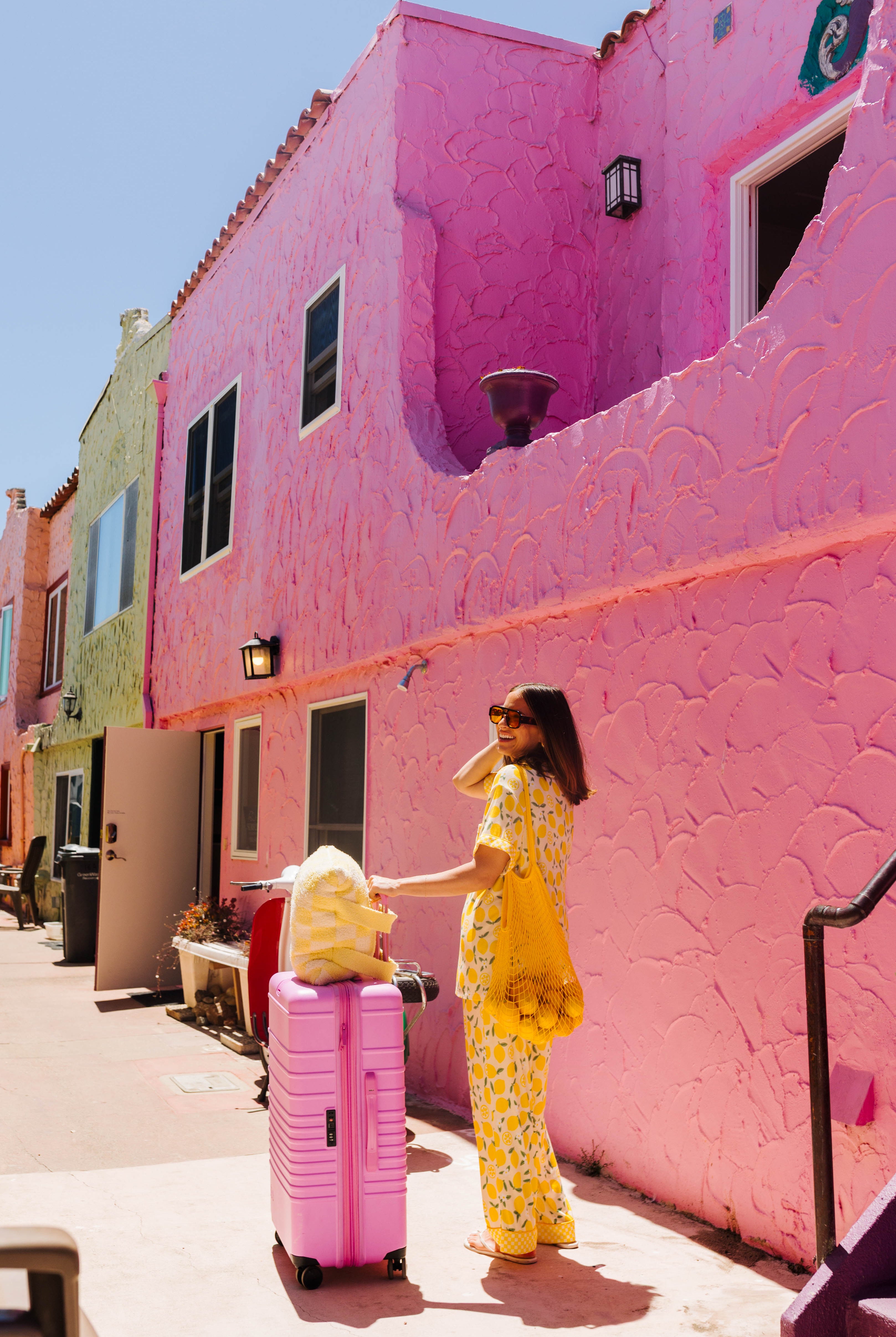 A woman in a yellow lemon pajama set stands next to a pink rolling suitcase on a sunny sidewalk beside a bright pink stucco building.