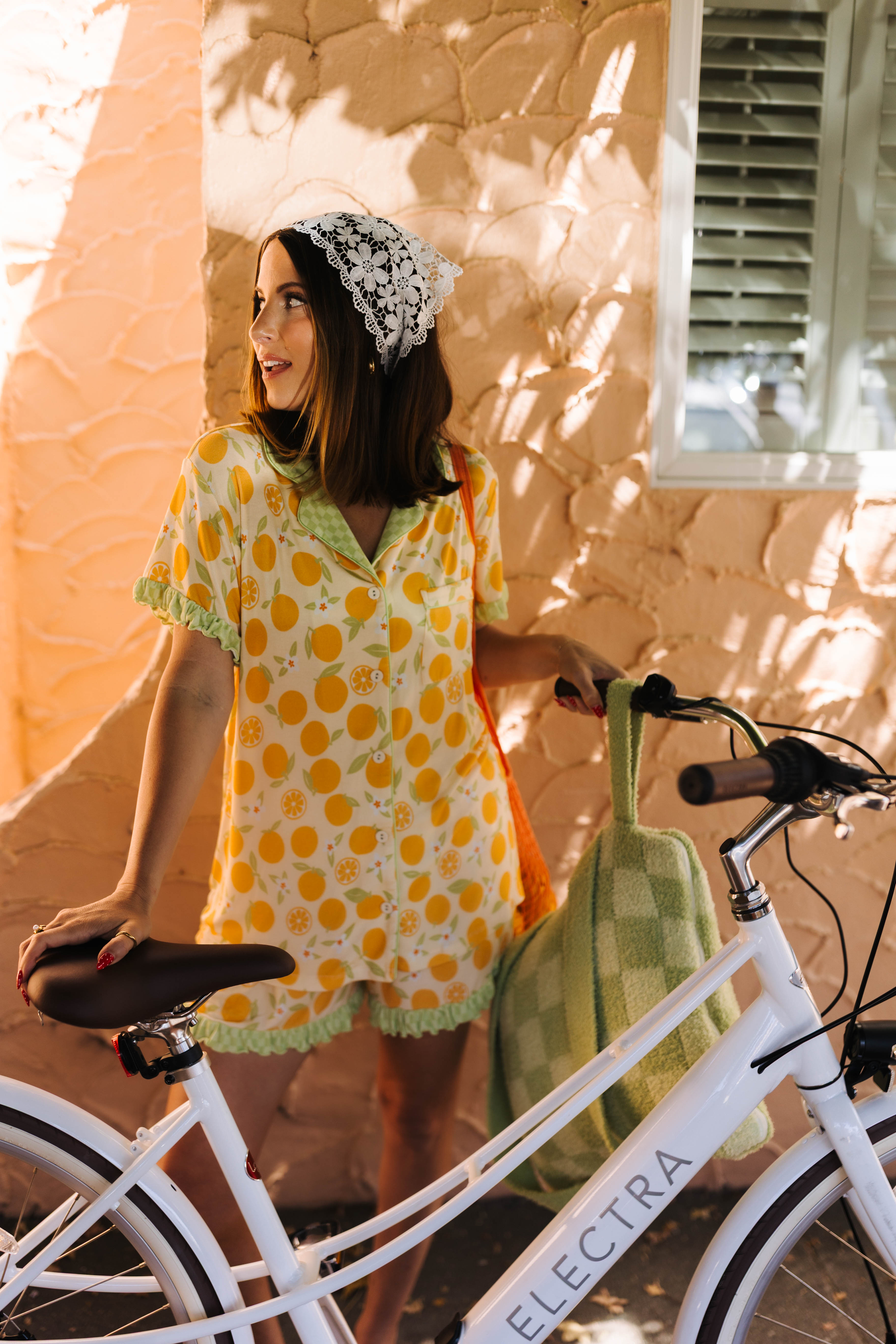 A woman wearing an orange patterned pj and headscarf stands next to a white Electra bicycle, holding a green checkered quillow against a textured stucco wall.
