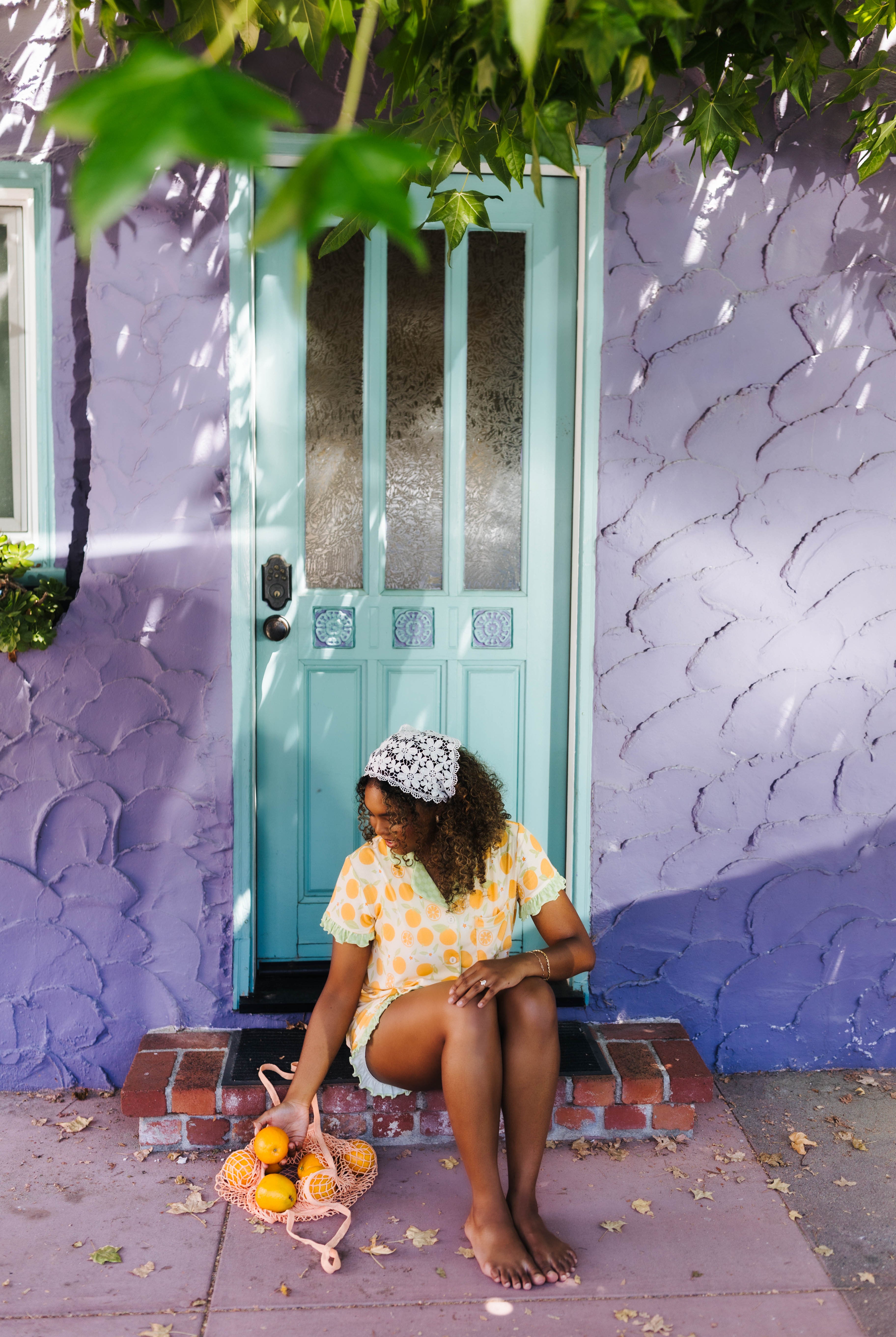 A woman sits barefoot on the steps of a purple stucco house wearing an orange pj set. She holds a small pink bag with fruit in front of a turquoise door.