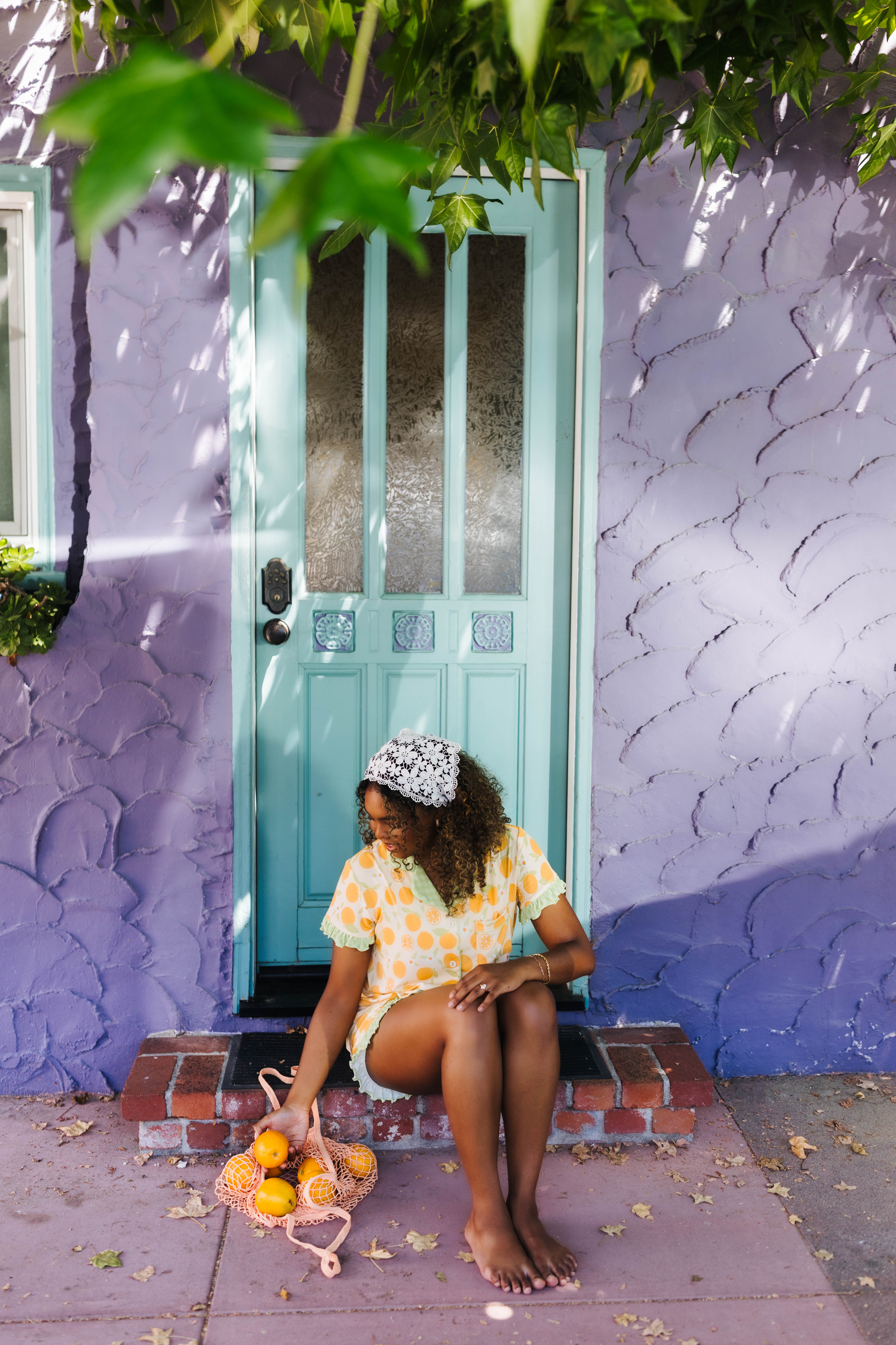 A woman sits barefoot on the steps of a purple stucco house wearing an orange pj set. She holds a small pink bag with fruit in front of a turquoise door.