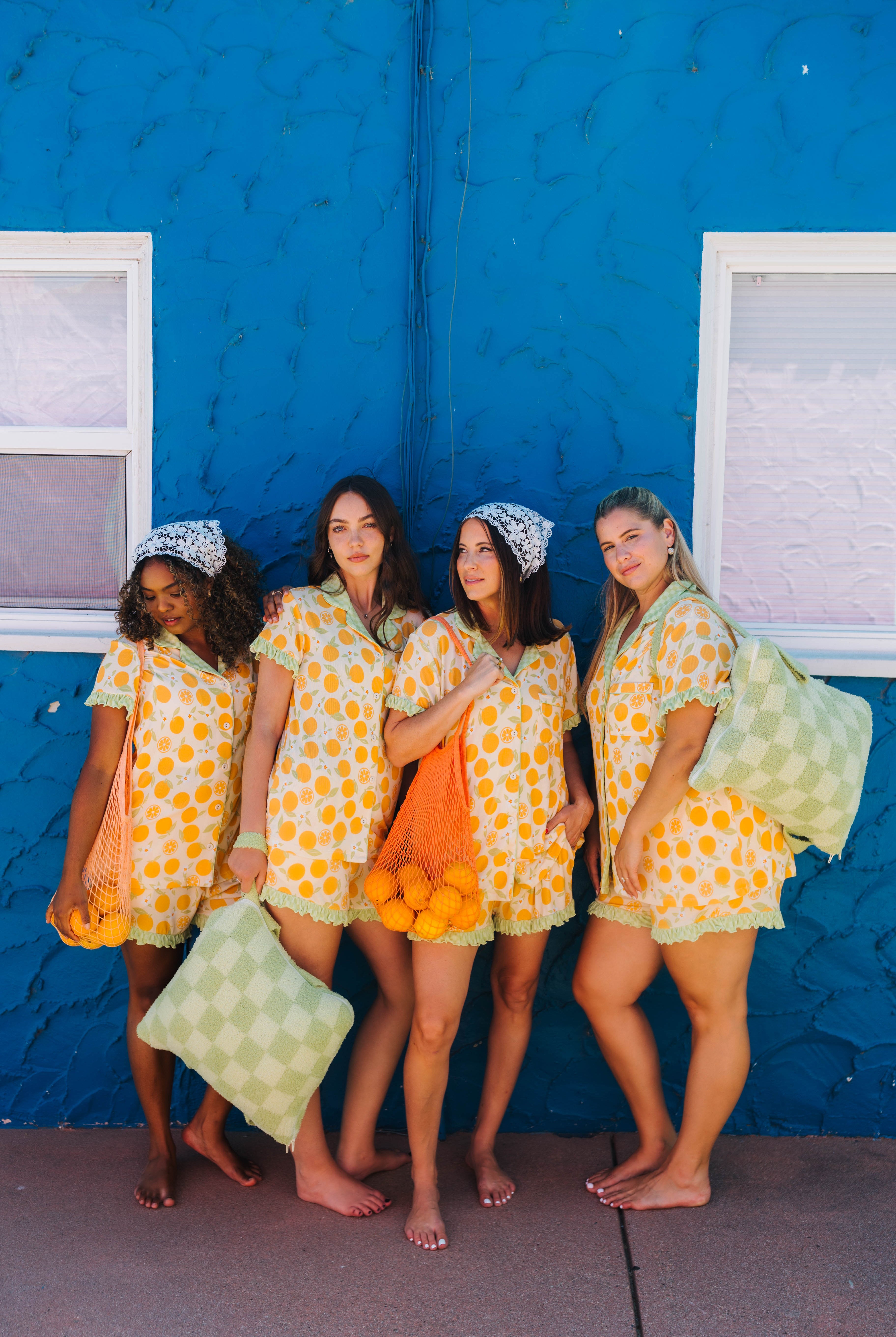 Four women posing together in matching yellow and orange patterned pjs standing barefoot against a bright blue stucco wall with windows.