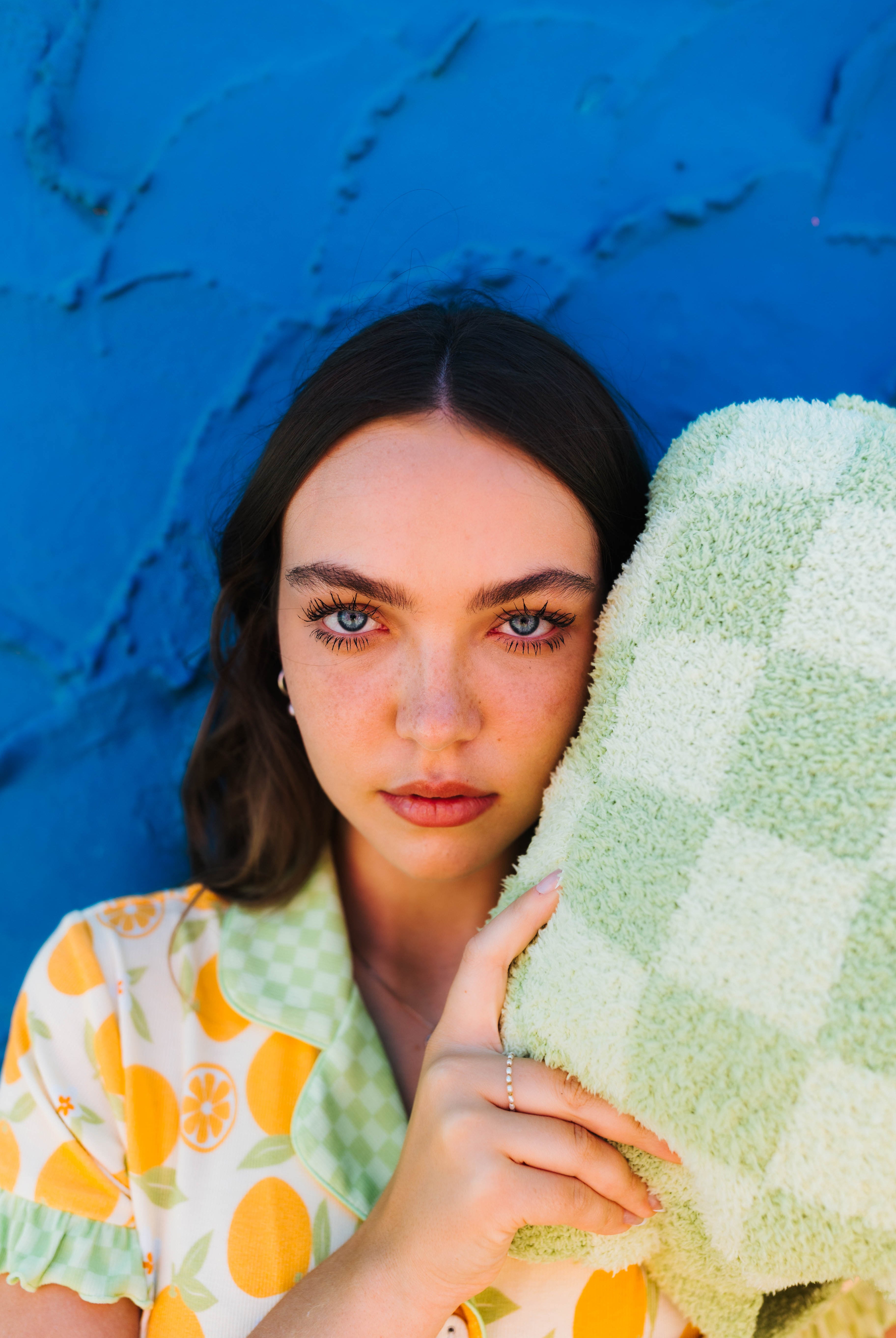 A woman with dark hair leans against a bright blue textured wall, holding a light green check quillow close to her face.