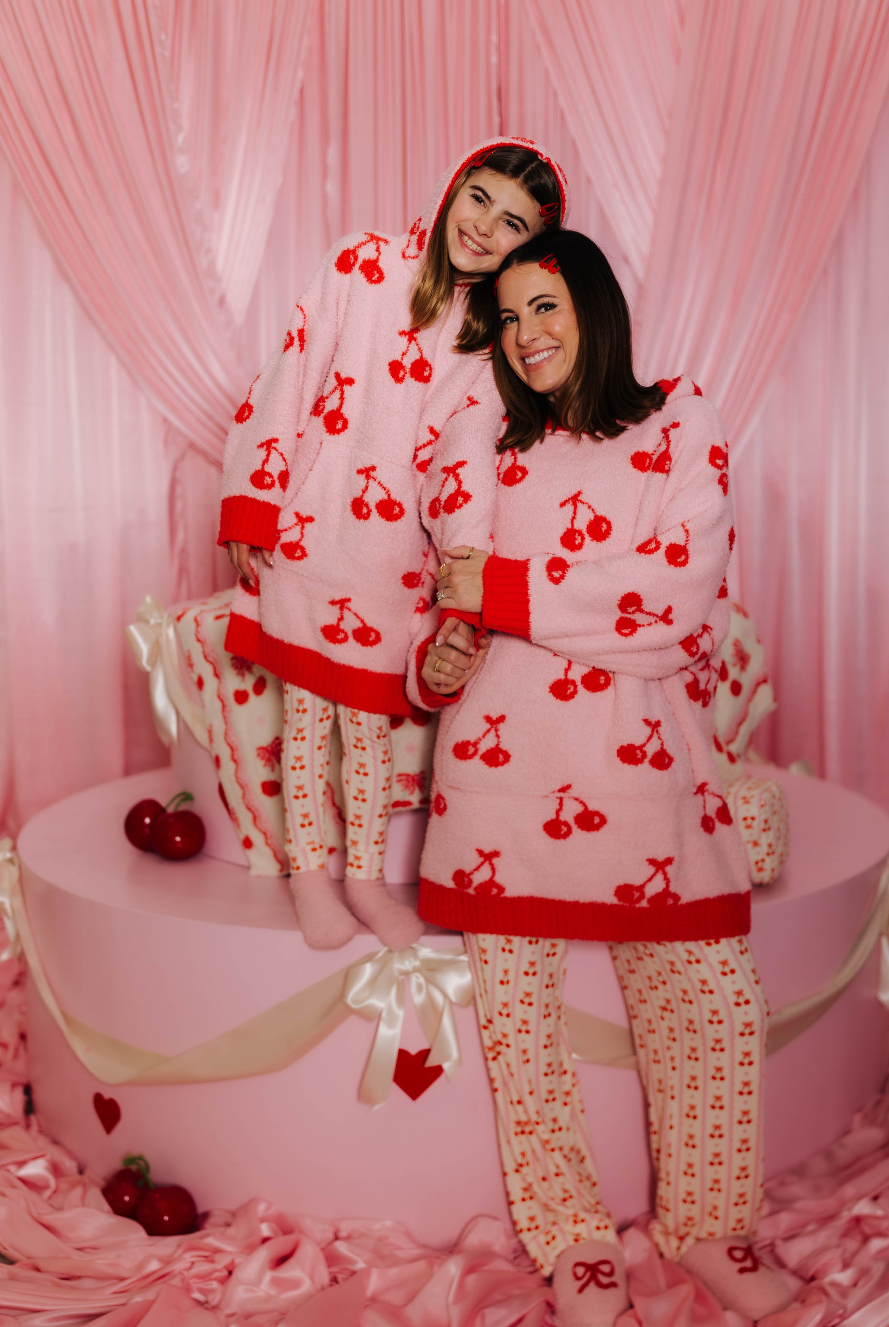 Krista and Kolly in matching cherry-patterned wearables standing in front of a pink curtain.