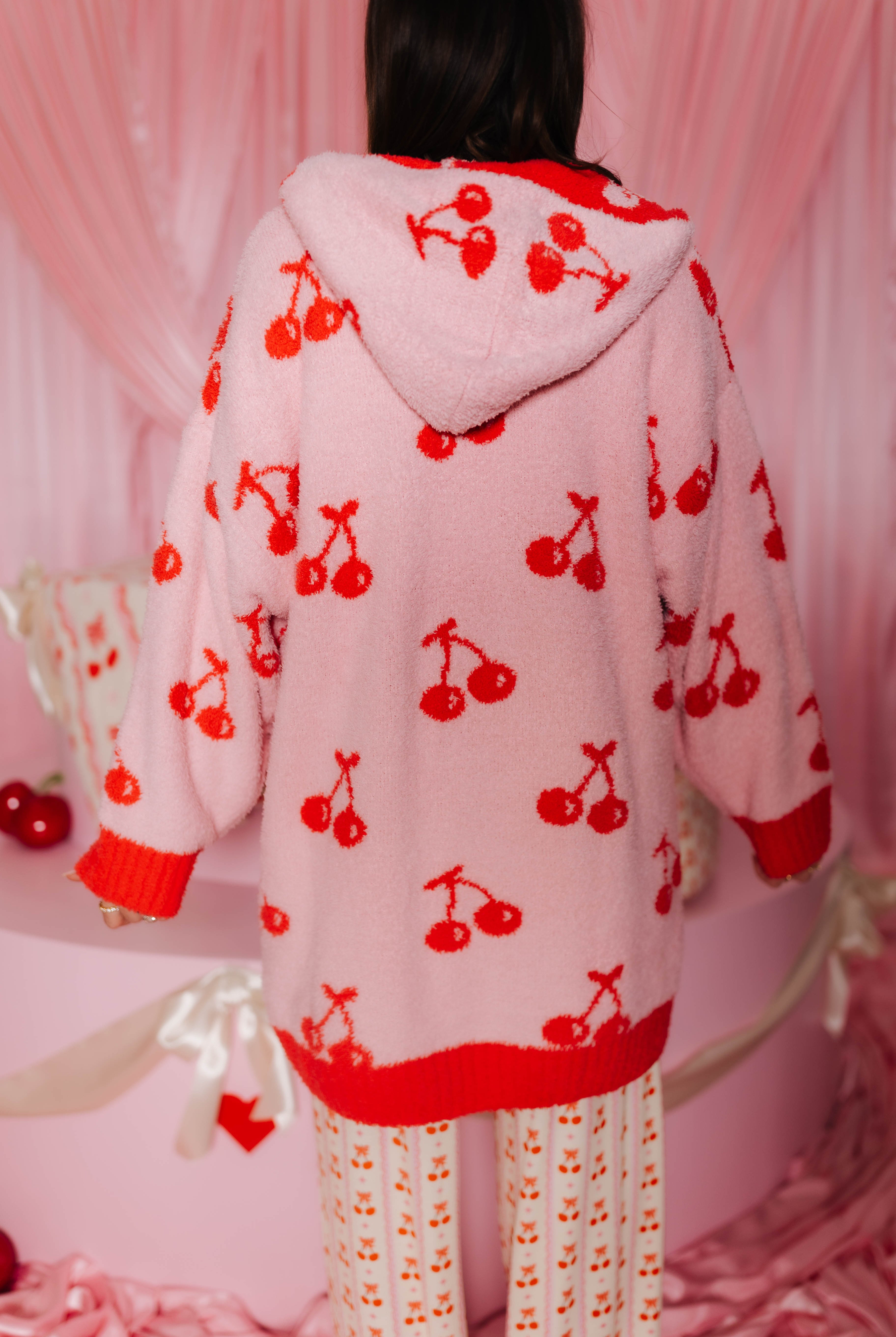 Person wearing a pink hoodie wearable with red cherry patterns in a pink-themed setting.