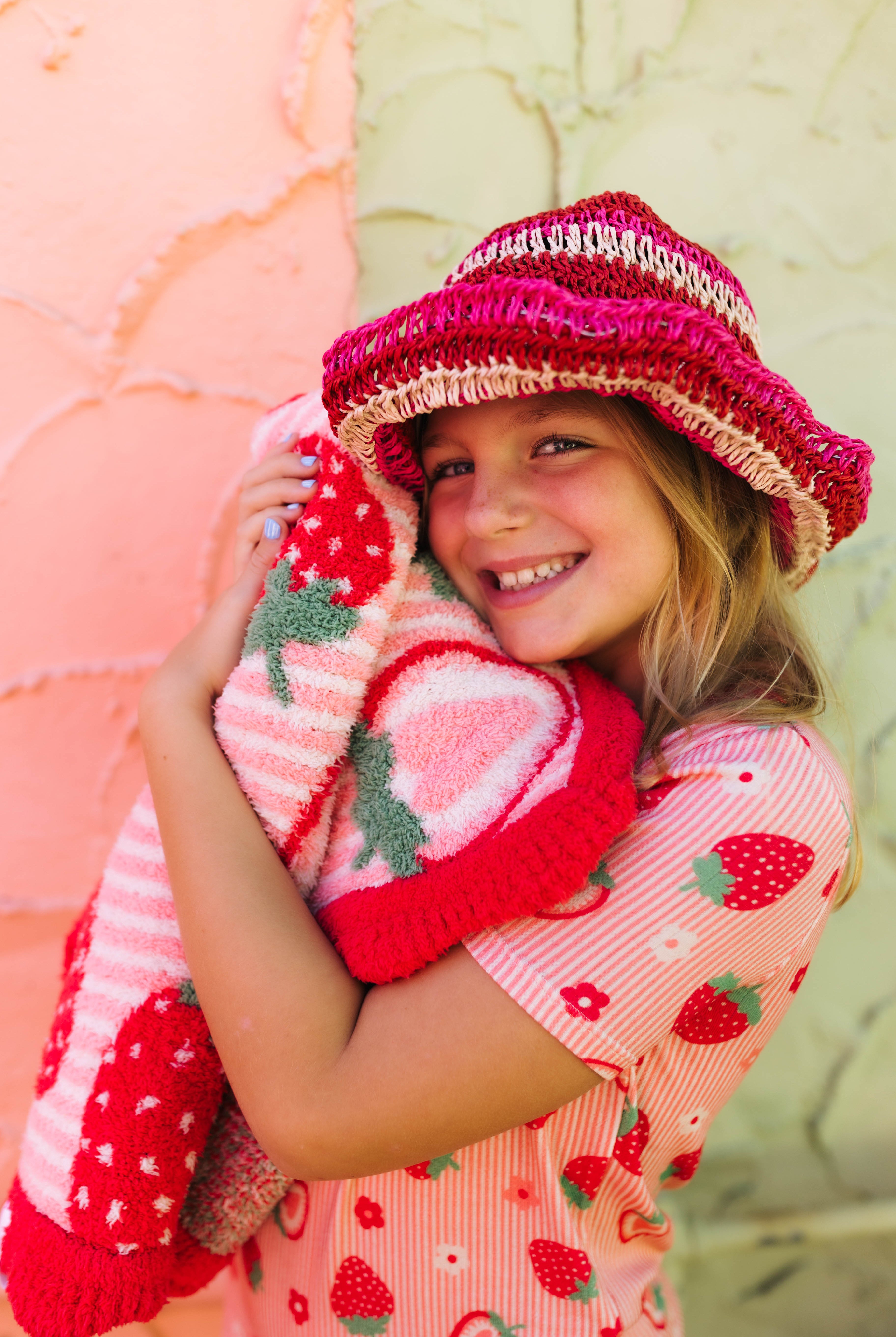 A smiling young girl wearing a pink and red strawberry-themed knit hat and matching strawberry-print top, hugging a pink and red strawberry knit blanket against a split peach and green textured wall.