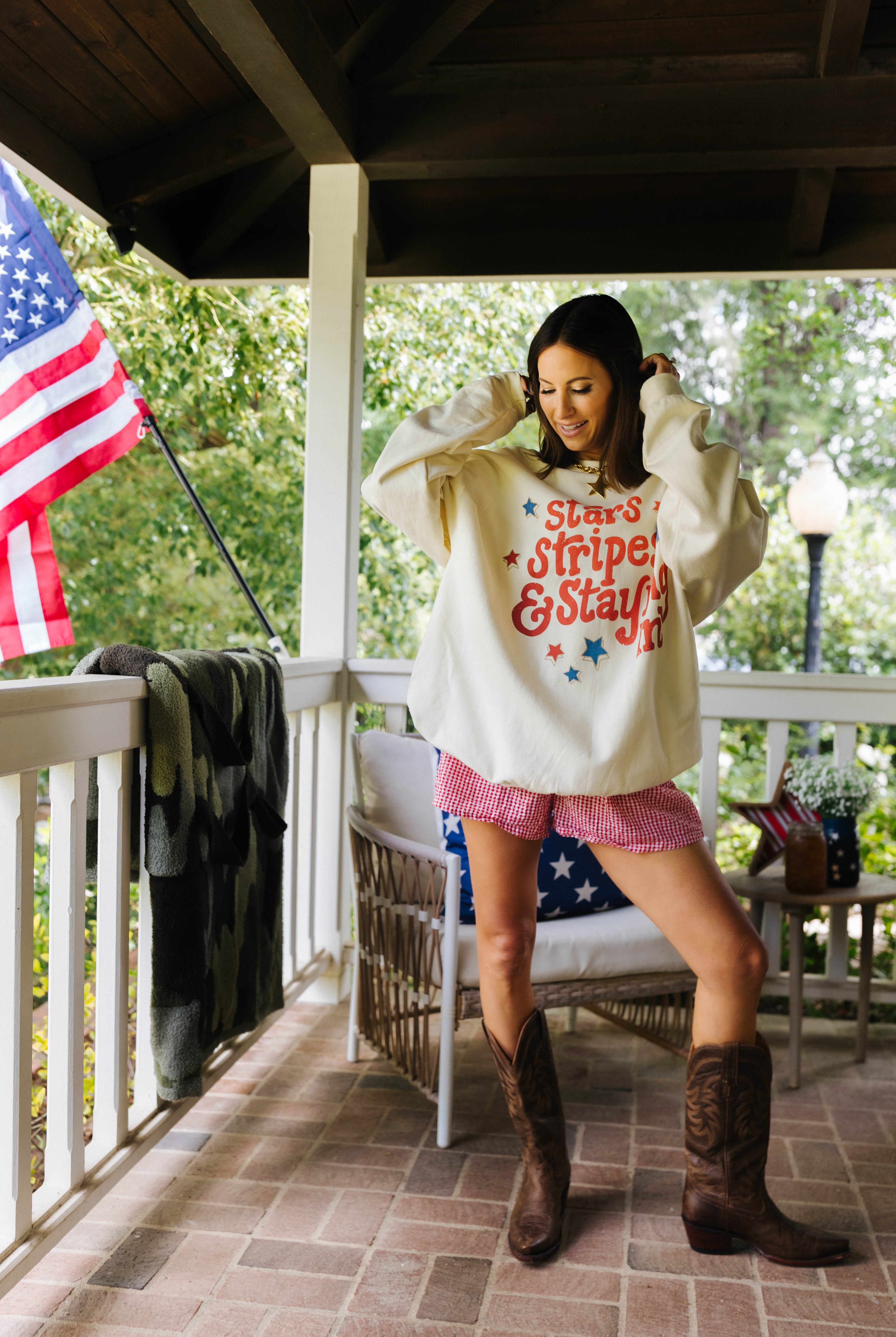 Woman on a porch wearing a sweatshirt with text and pink shorts, standing next to an American flag.