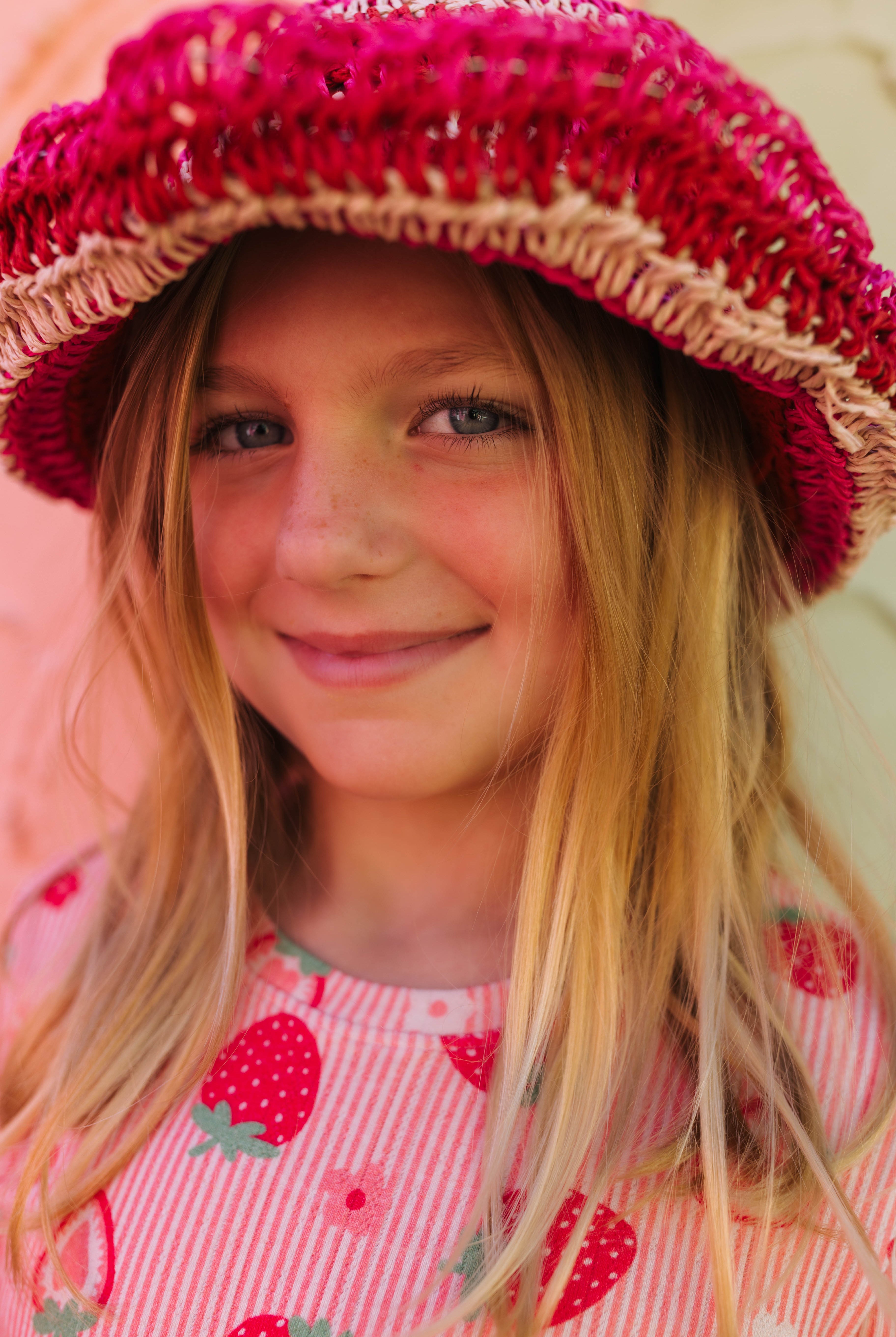 A young girl with long blonde hair smiling while wearing a bright pink and red striped knit hat with a straw-like trim and a pink strawberry-patterned top.