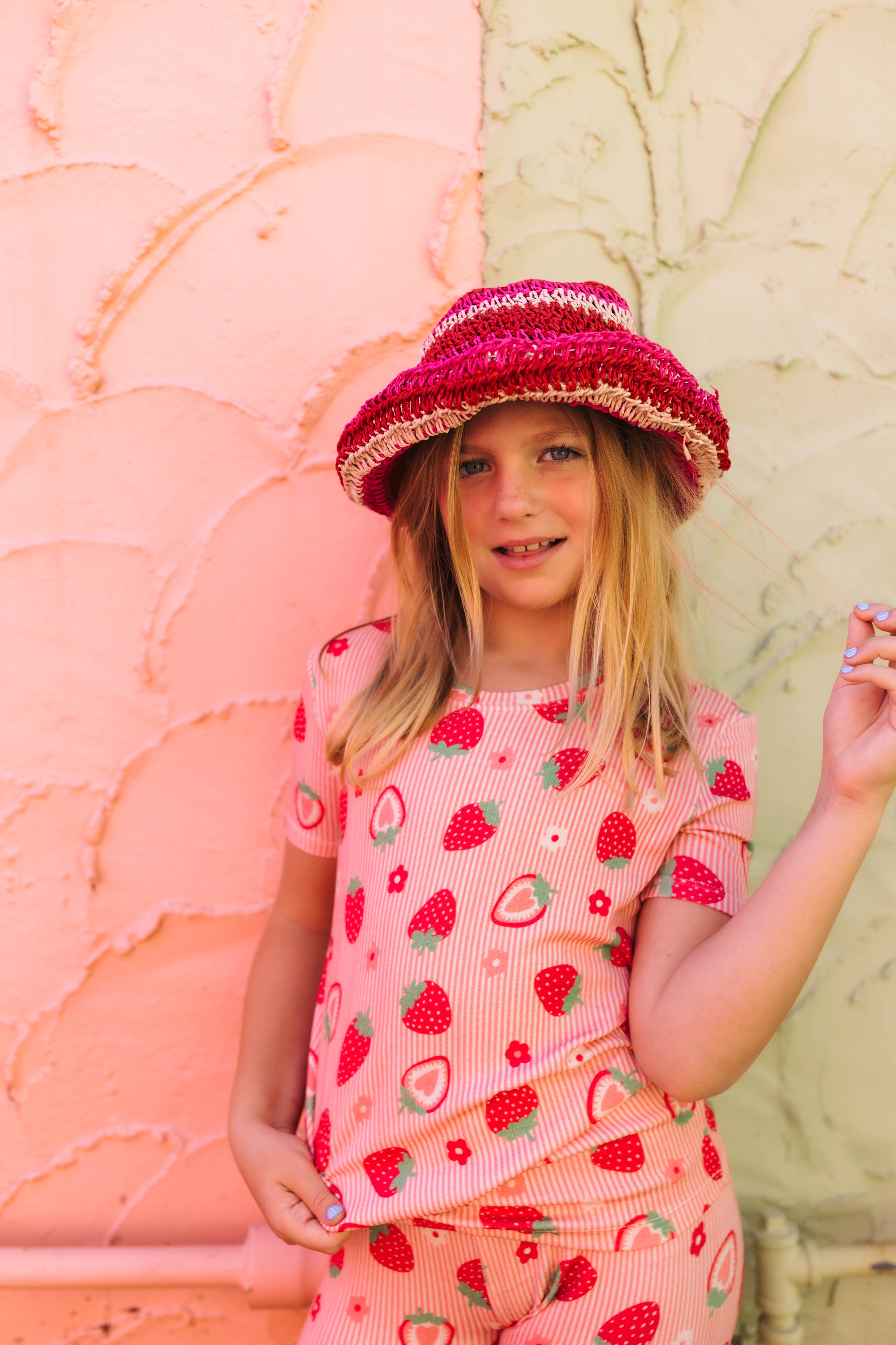 A young girl wearing a pink strawberry-print pajama set and a matching pink knit sun hat, standing against a split pink and green stucco wall.