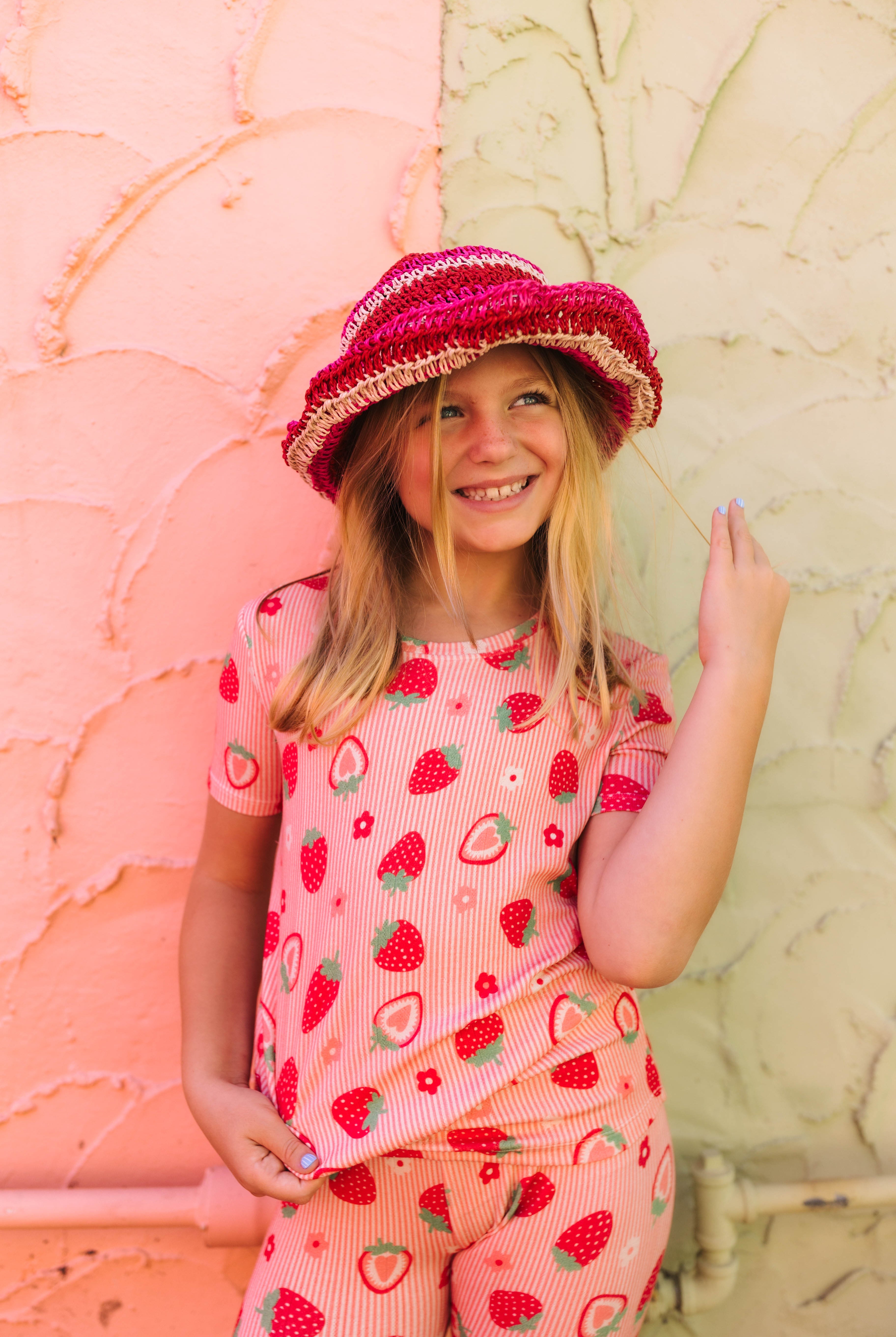 A young girl smiling while wearing a pink strawberry-patterned short-sleeve top and matching pants, along with a pink woven hat, standing against a split pink and beige textured wall.