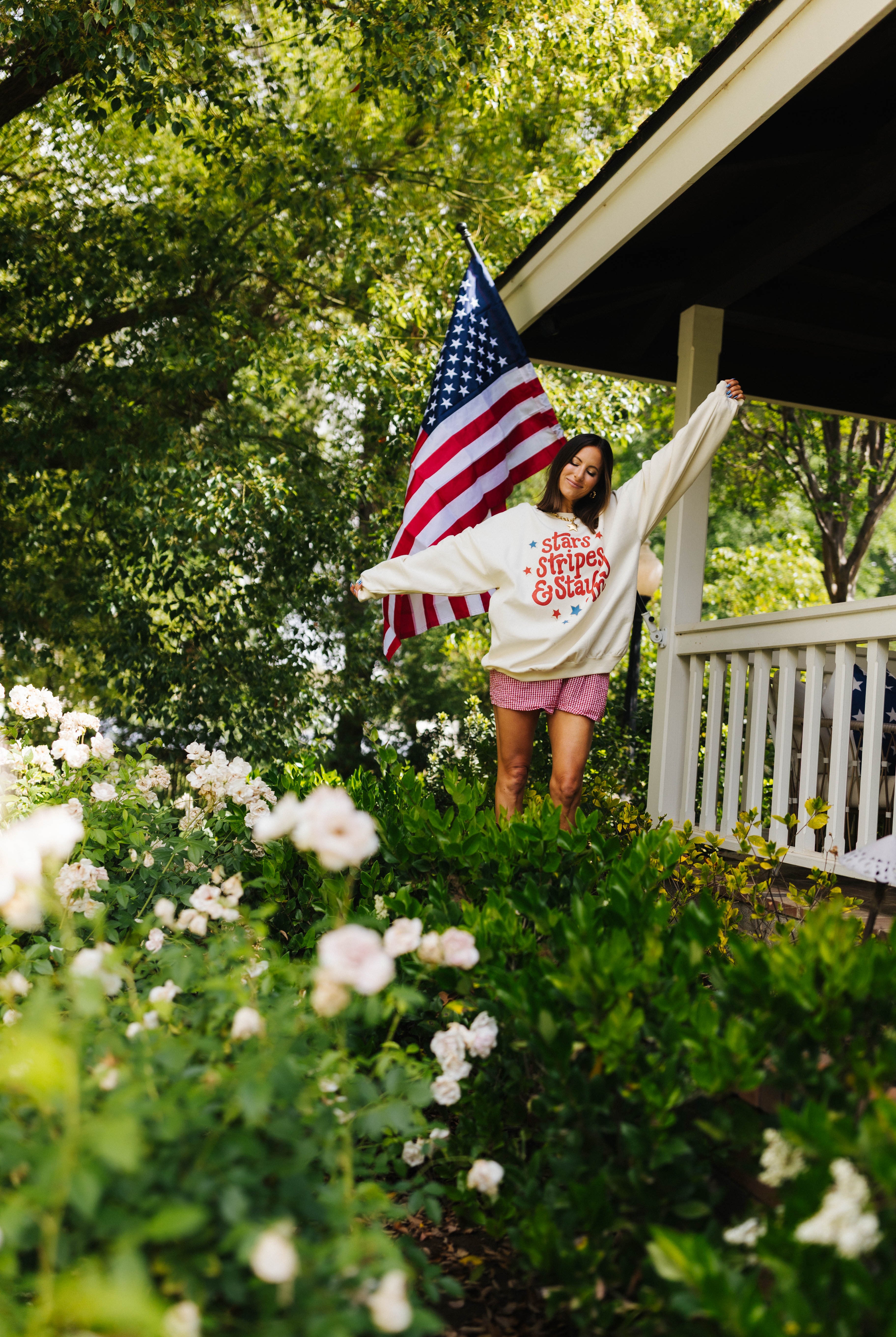 Woman standing in front of American flag on a porch with greenery and flowers