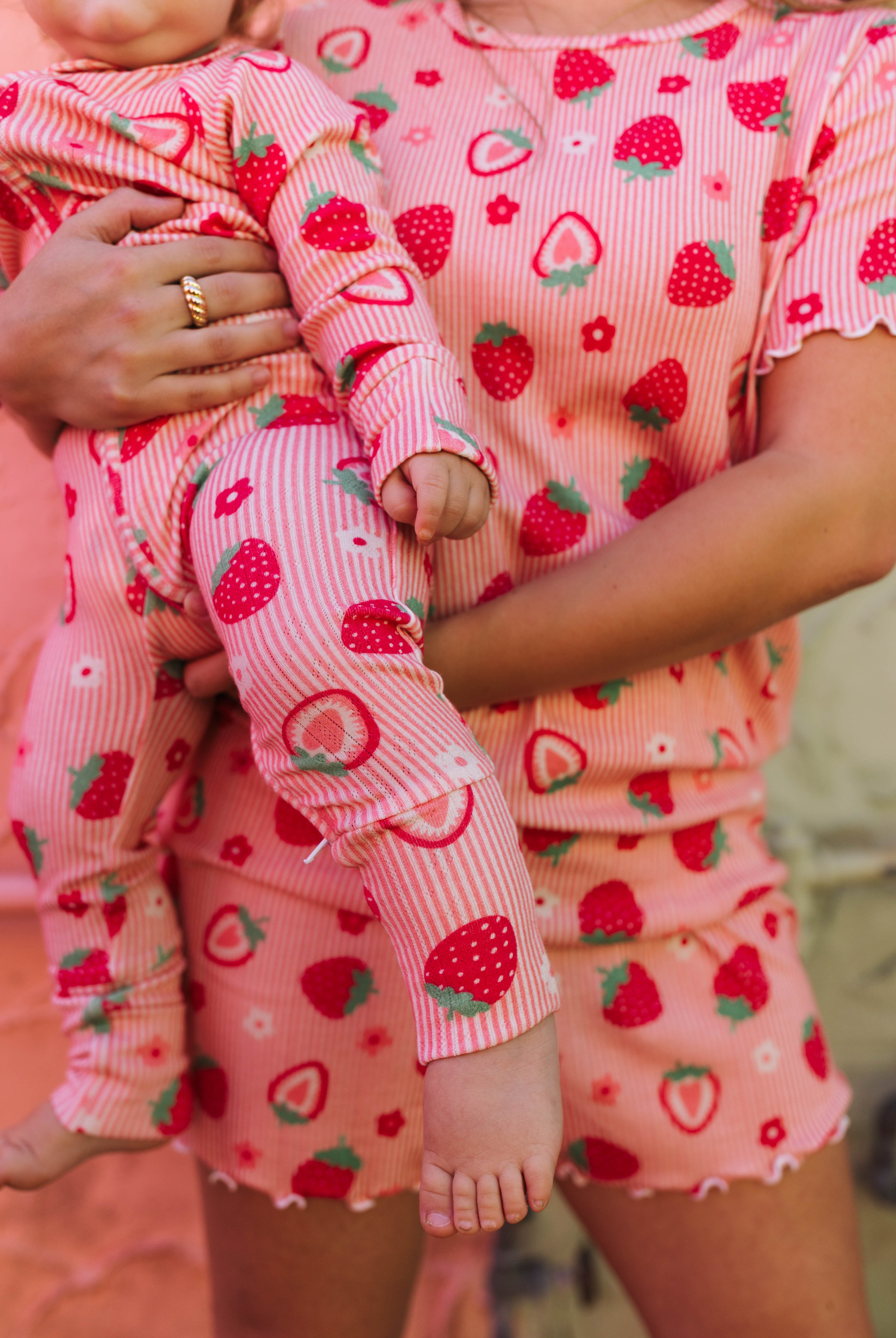 A close-up lifestyle shot of a mother and child wearing matching strawberry print pajamas. The set includes a short-sleeve top and shorts for the adult, and a long-sleeve pajama set for the baby, both featuring a pink and white striped base with red strawberries and green leaves.