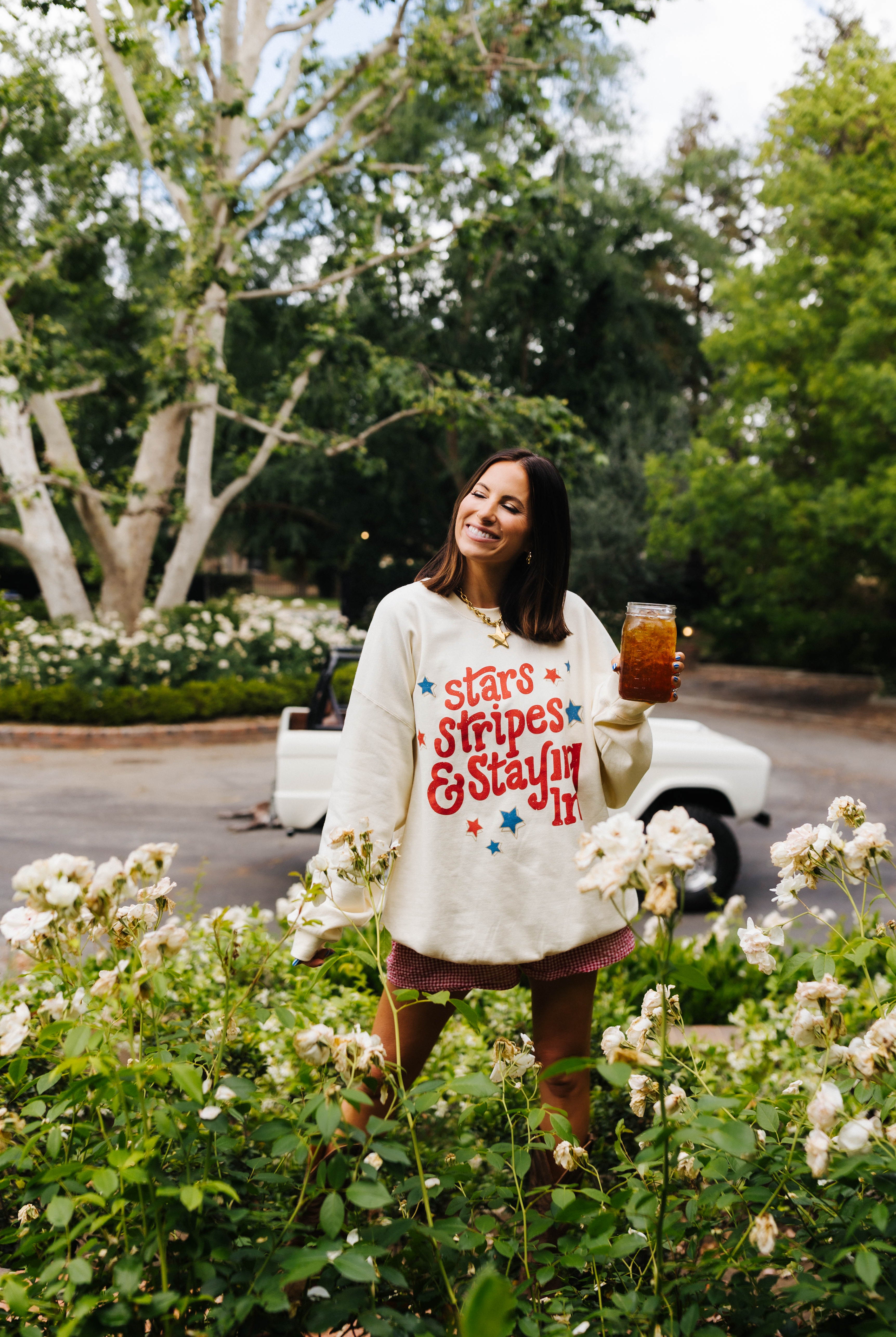 Woman in a garden wearing a 'Stars & Stripes & Stayin' It' shirt, holding a drink.