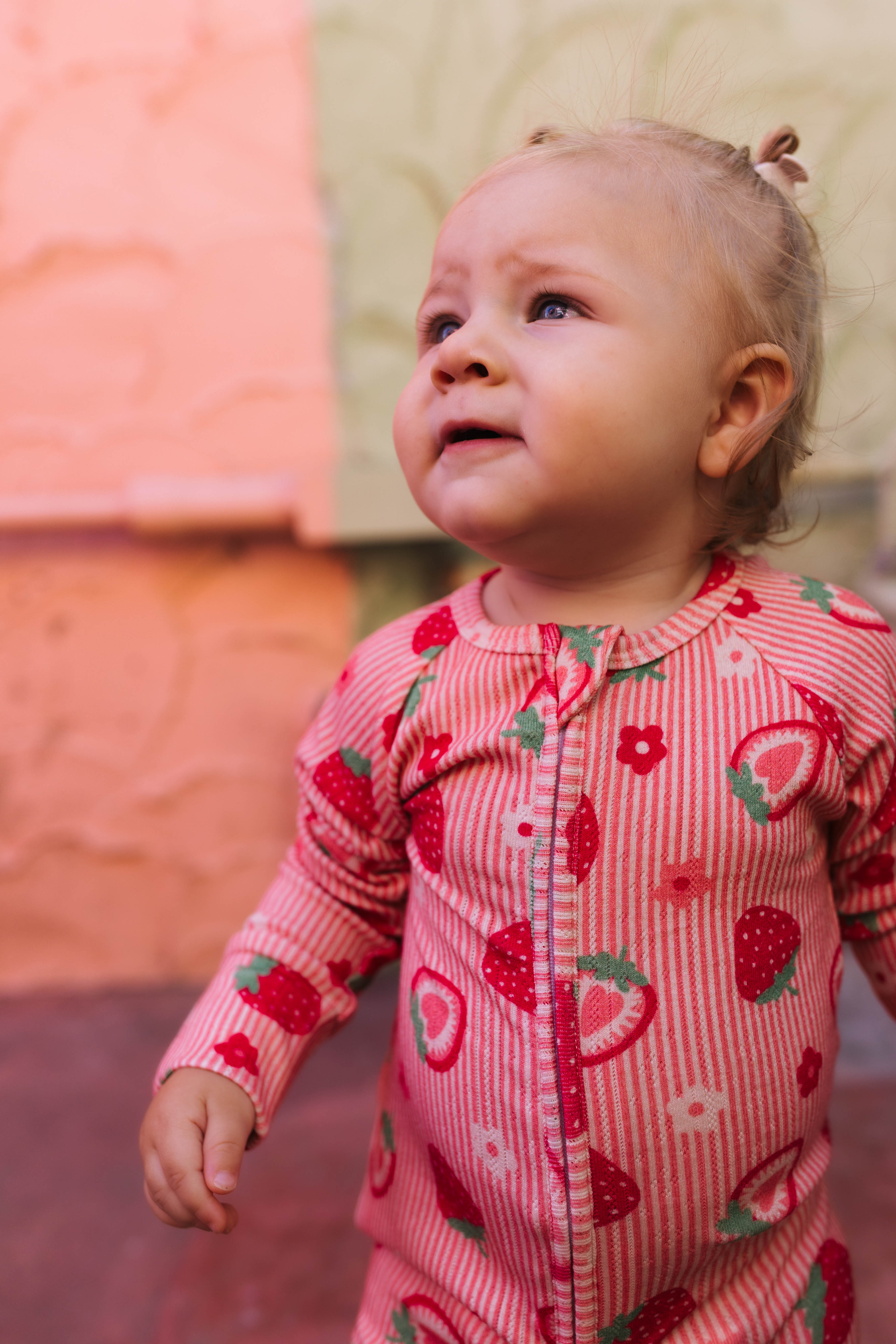 A baby girl wearing a pink and white striped long-sleeve romper with a strawberry and flower print, looking upward outdoors.