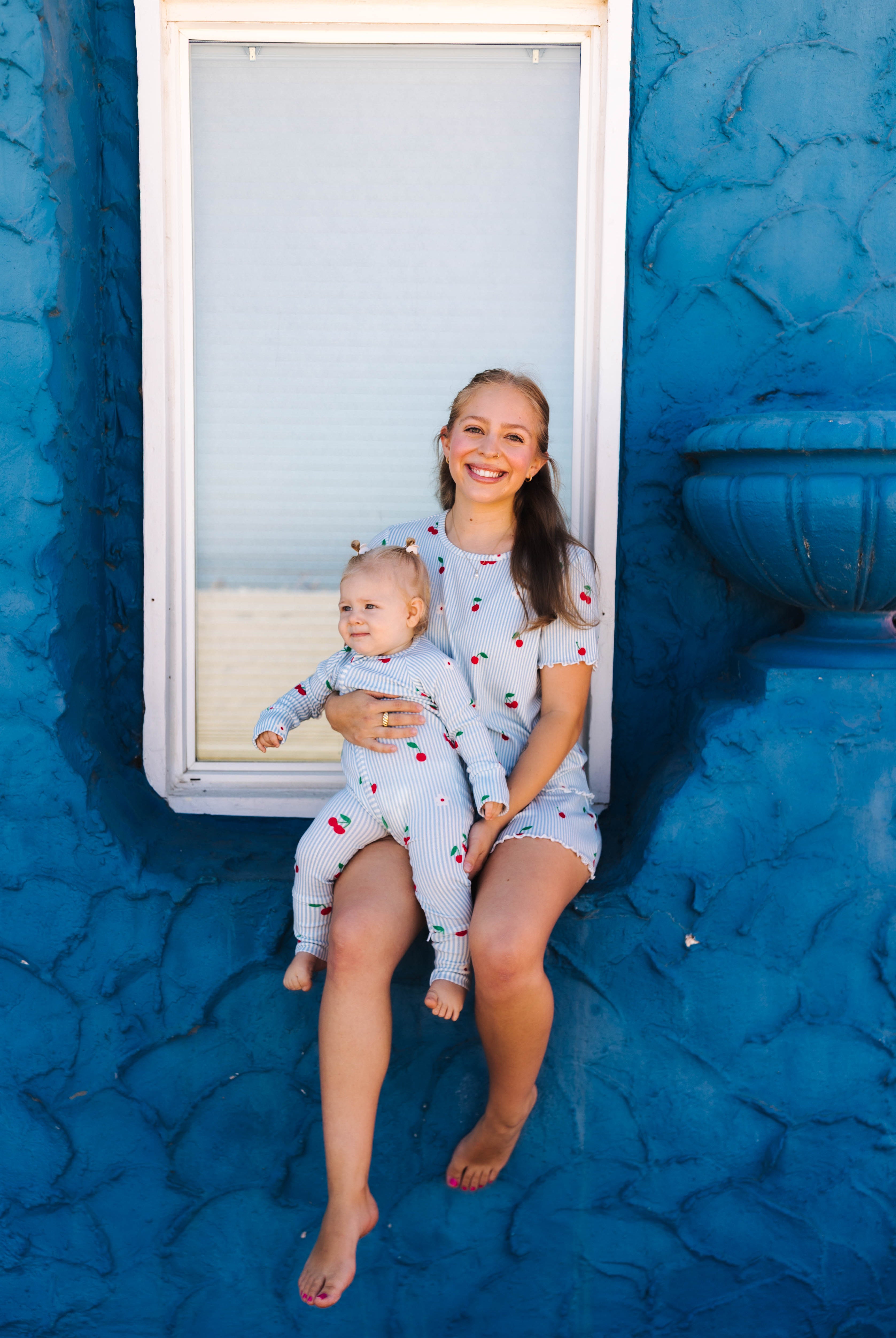 A smiling woman sits barefoot on a textured blue wall holding a baby. Both are wearing matching light blue pajamas with a cherry pattern.