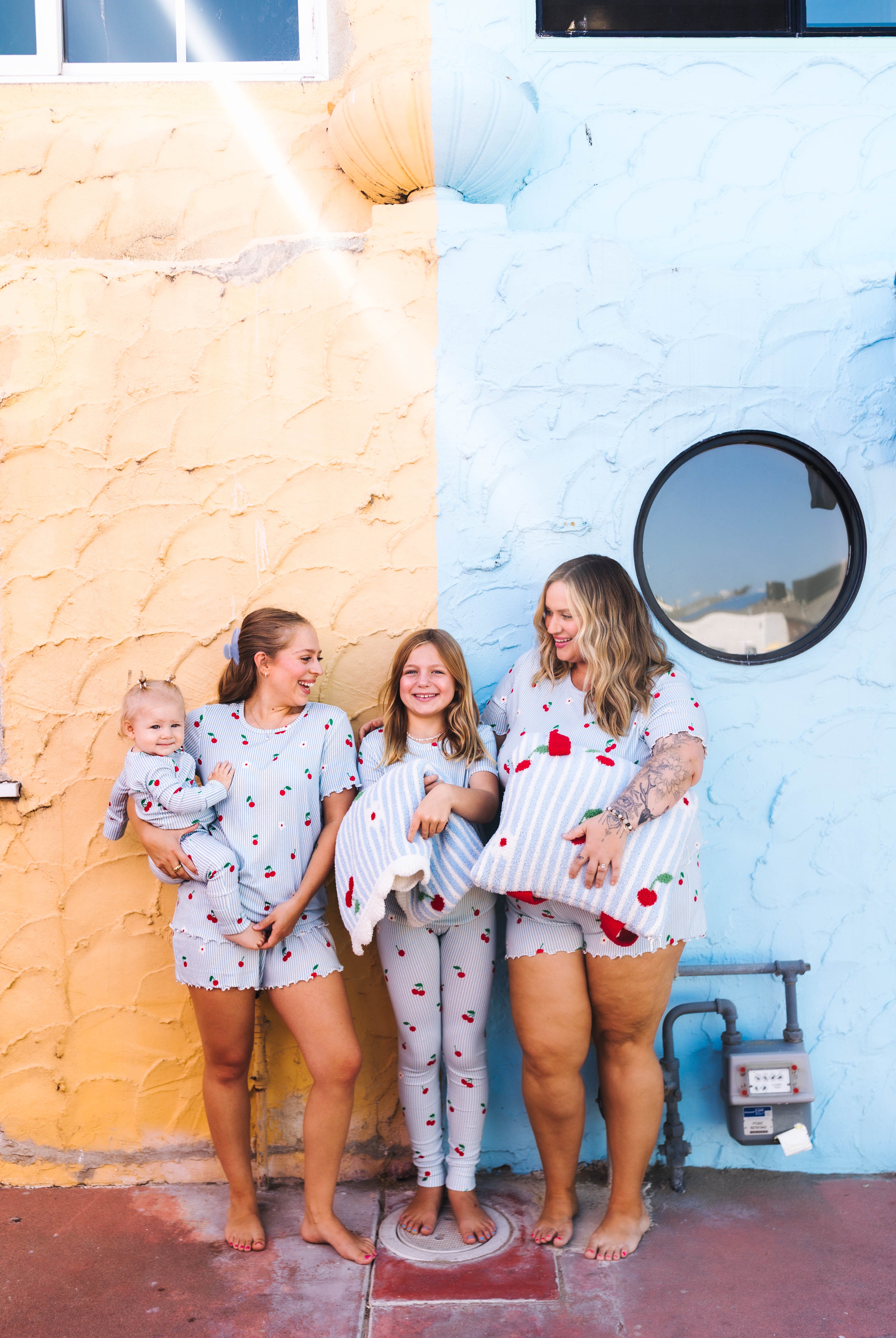Four girls wearing matching strawberry-themed pajamas stand barefoot outdoors against a split yellow and light blue stucco wall all in matching cherry pajamas!