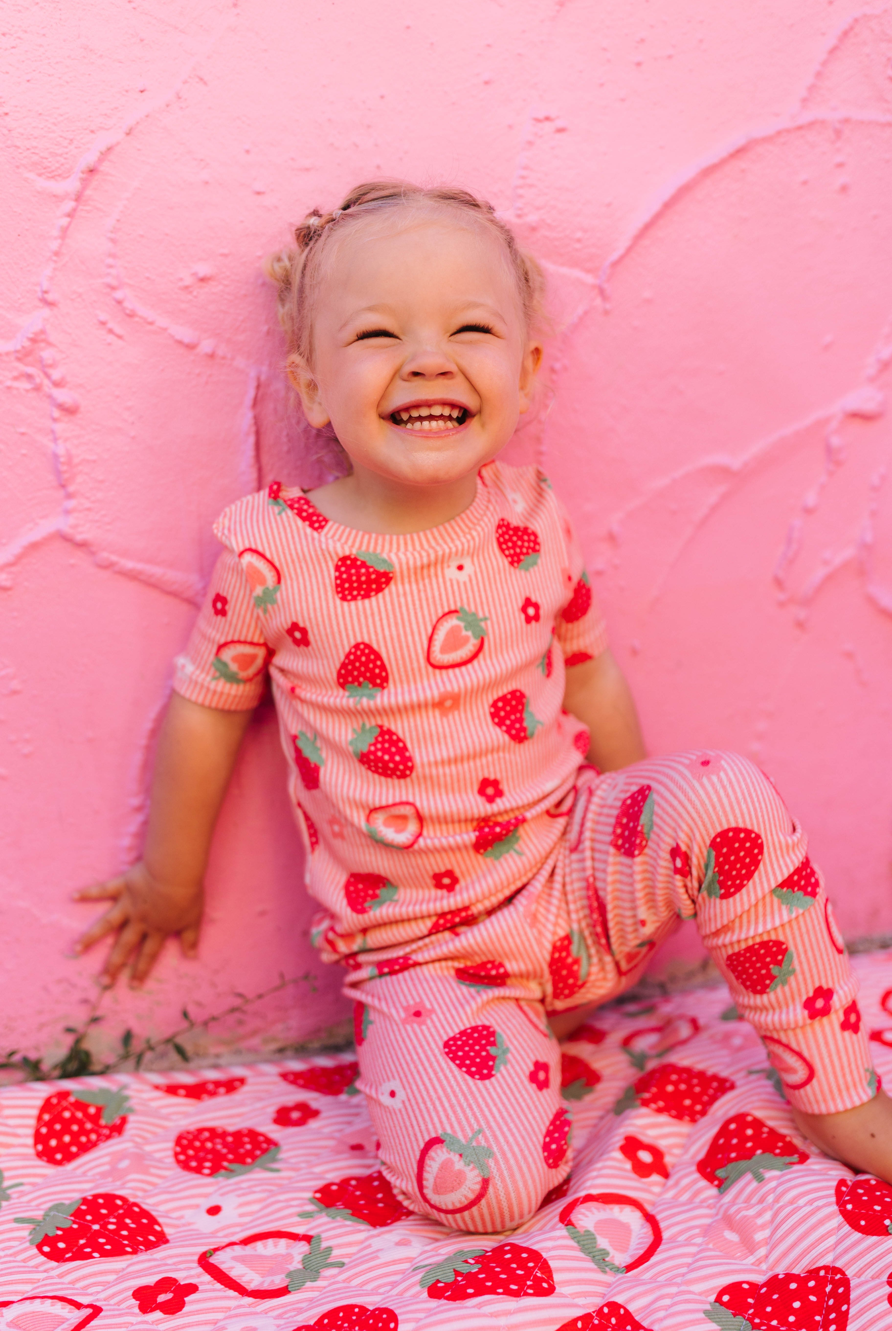 A smiling toddler wearing a pink strawberry-print pajama set sits on a matching strawberry-patterned blanket against a pink textured wall.