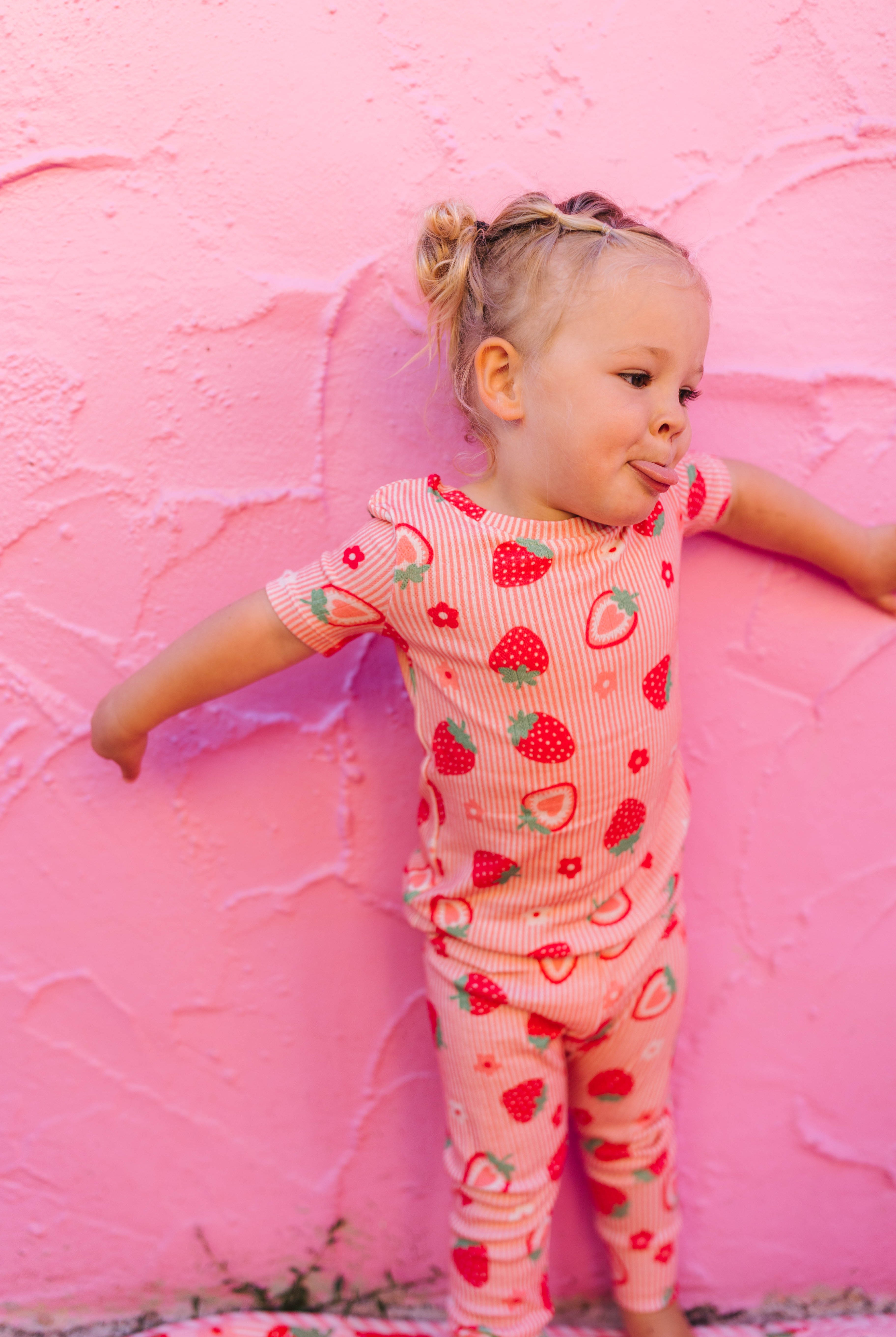A toddler girl wearing a pink and red strawberry print short-sleeve pajama set, posing against a textured pink wall.