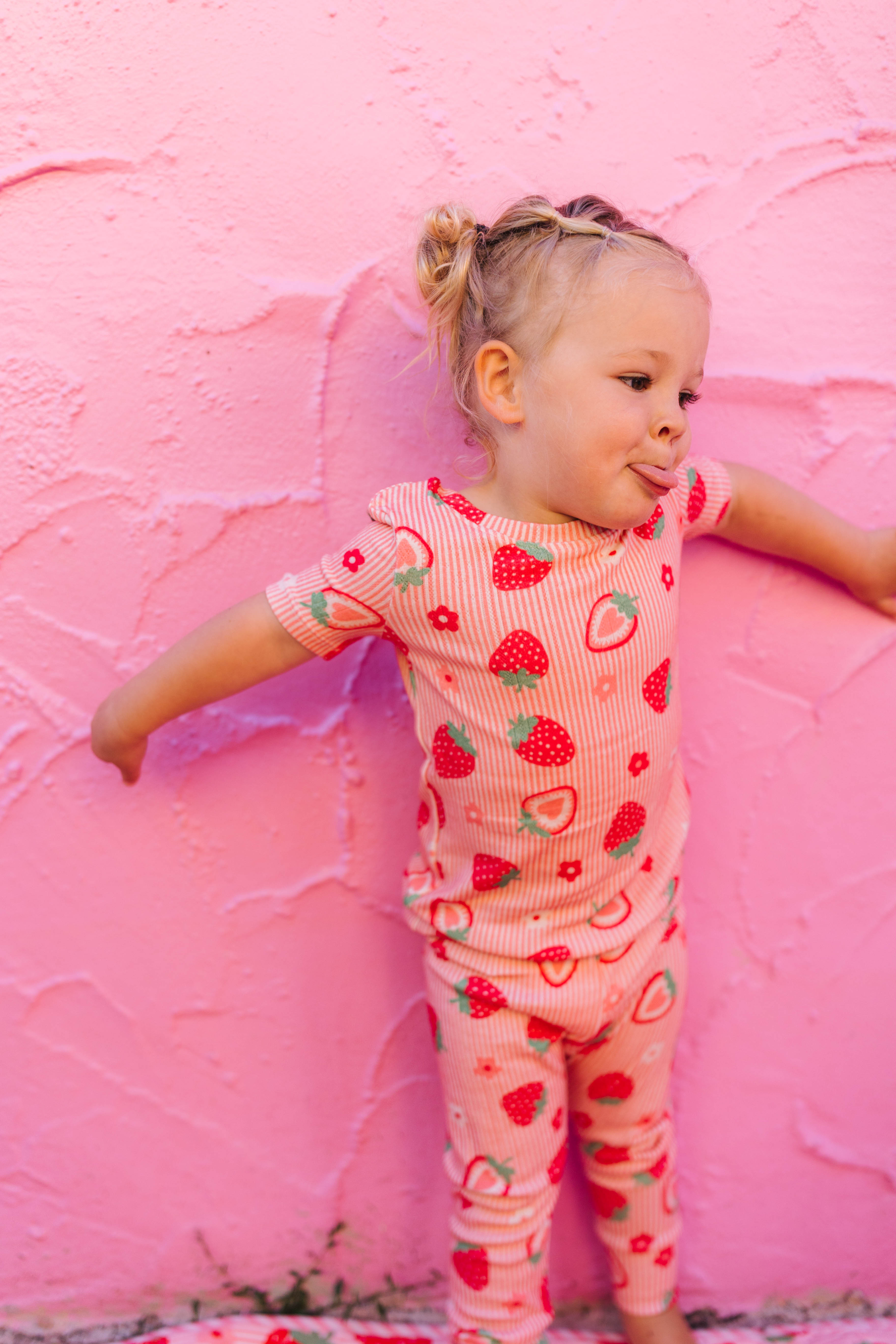 A toddler girl wearing a pink and red strawberry print short-sleeve pajama set, posing against a textured pink wall.