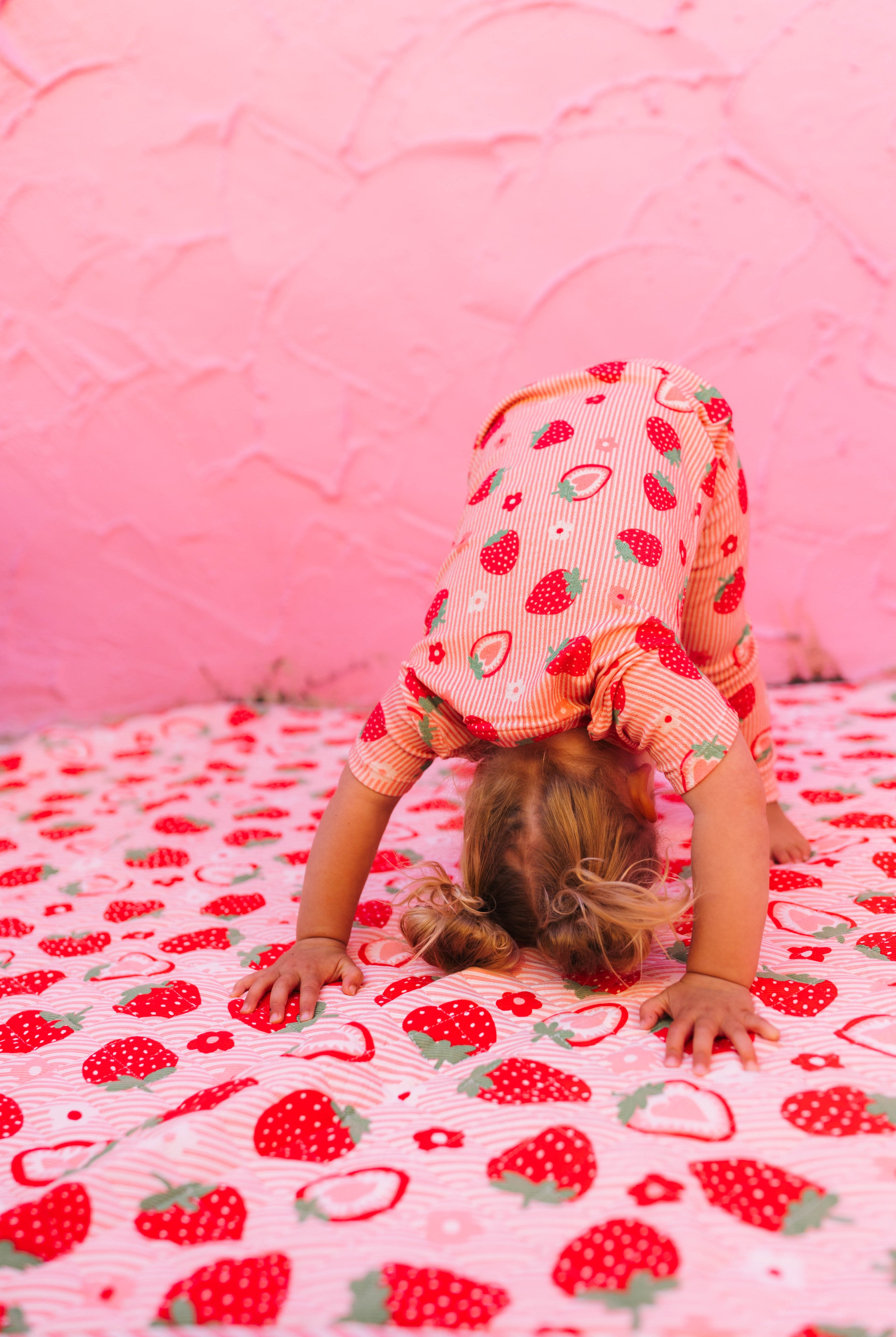 A young child wearing a pink and red strawberry-patterned pajama set is doing a headstand on a matching strawberry-print blanket against a pink textured wall.