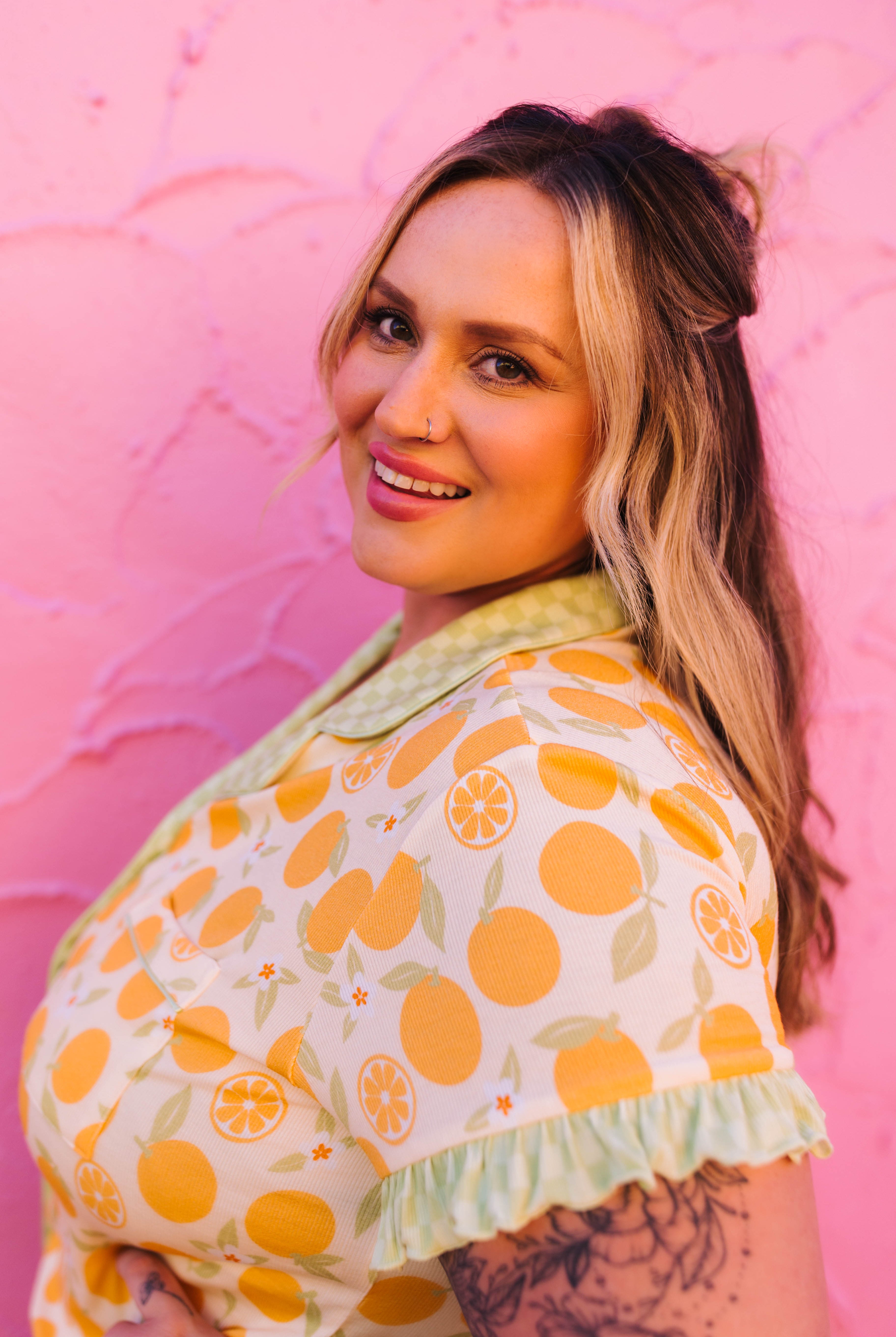A woman with blonde-highlighted hair smiling and looking over her shoulder, wearing a short-sleeve pj top with a yellow-orange citrus fruit print and a pale green ruffled collar and hem, posed against a textured pink wall.
