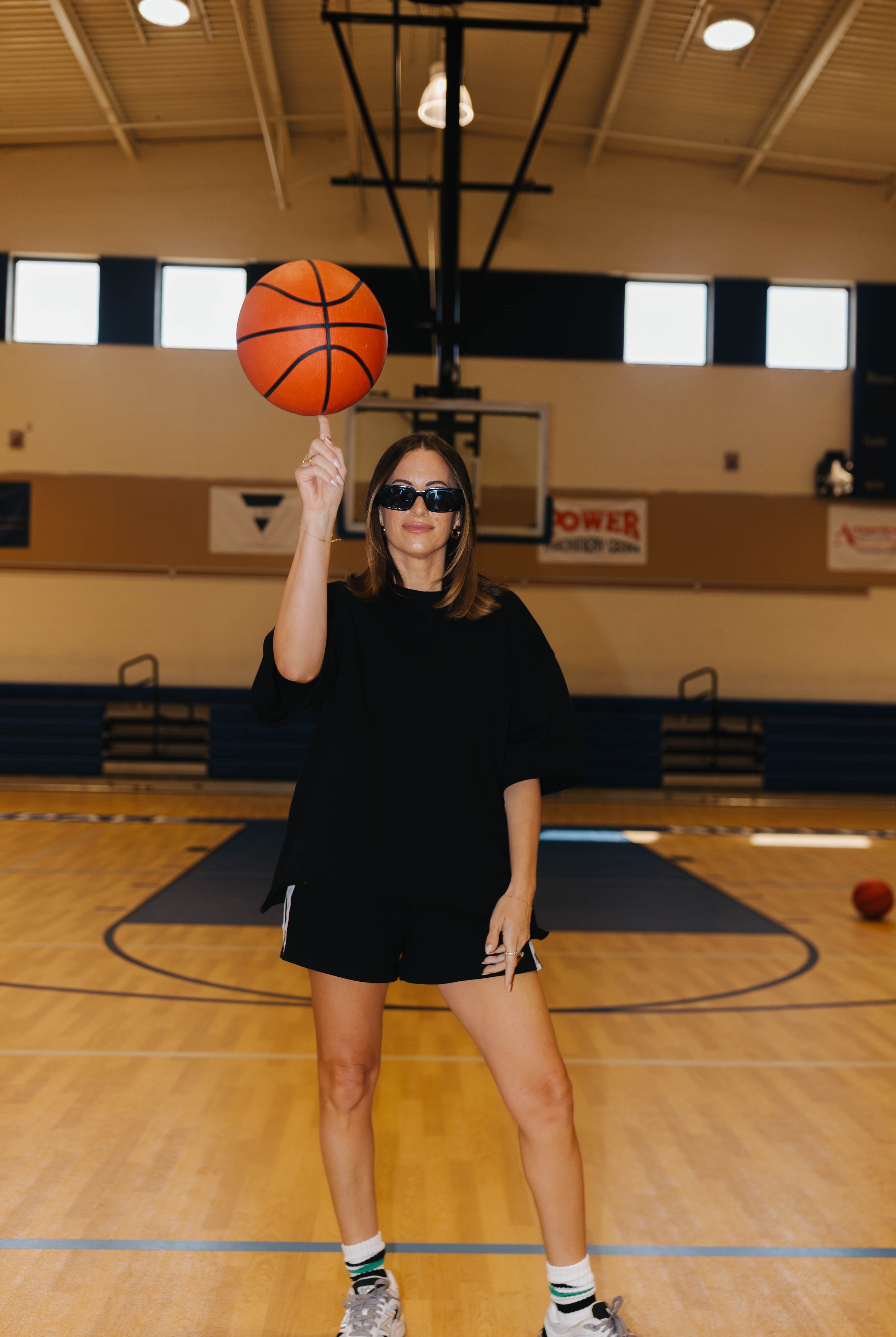 A woman wearing a black oversized t-shirt and black shorts stands on a basketball court, balancing an orange basketball on her finger. She is wearing sunglasses, white socks, and sneakers.