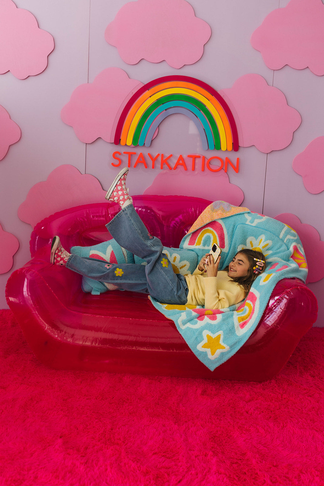 Person lying on a pink inflatable chair with a rainbow and 'STAYKATION' text against a pink cloud-patterned background.