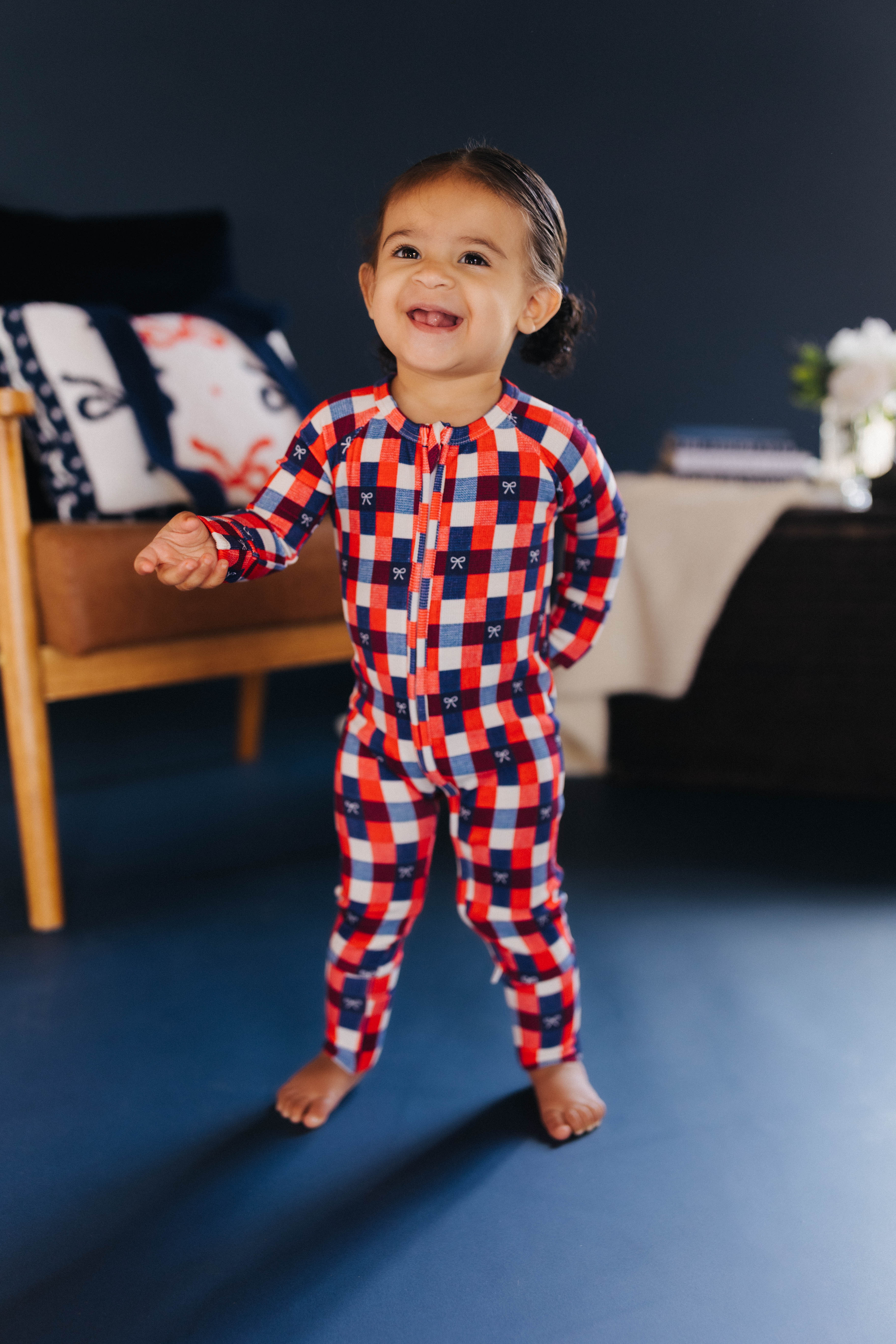 Child wearing a red, blue, and white gingham onesie in a room with a couch and pillows.