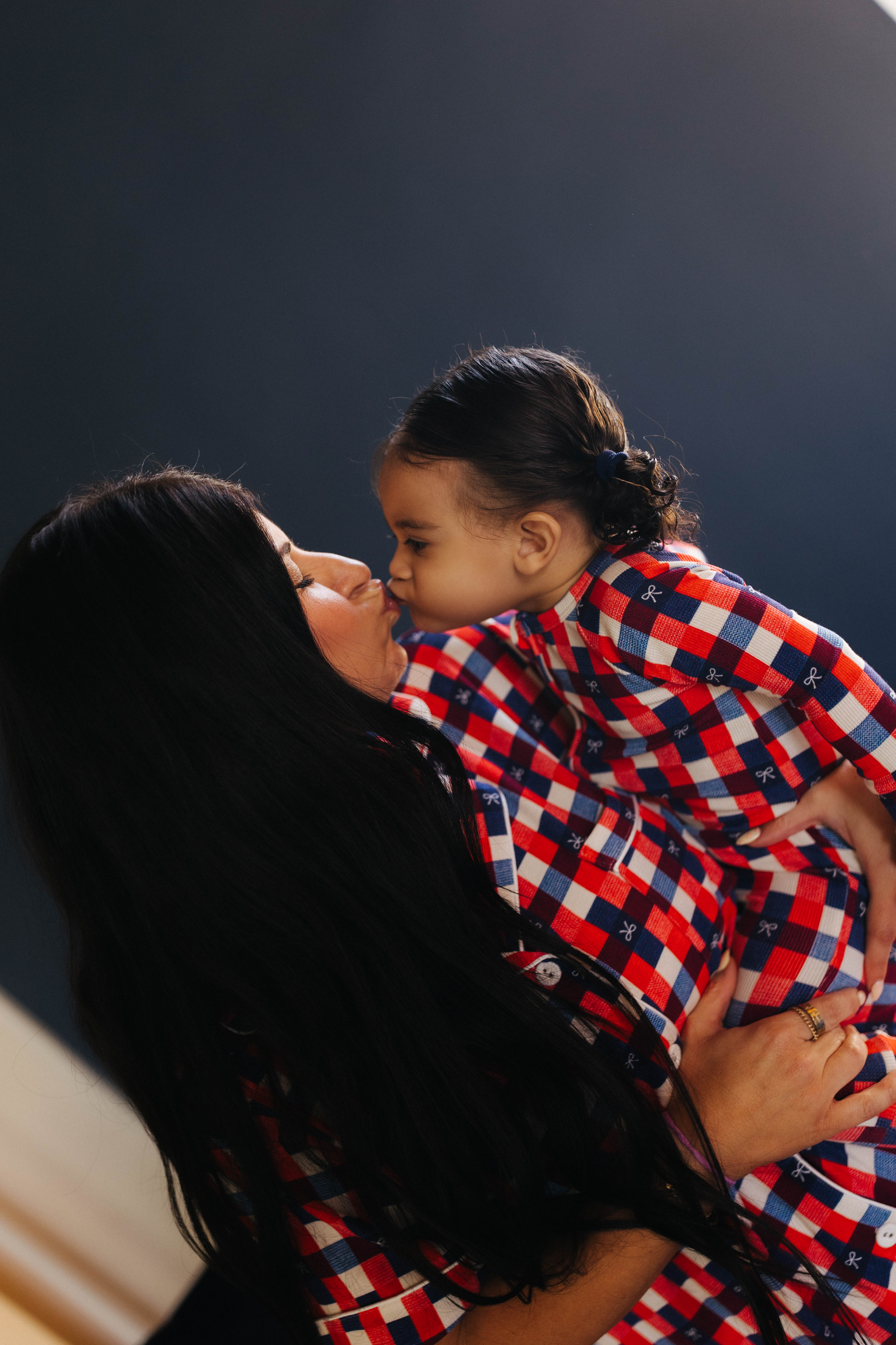 Woman holding a child in a red, white, and blue gingham outfit against a dark background