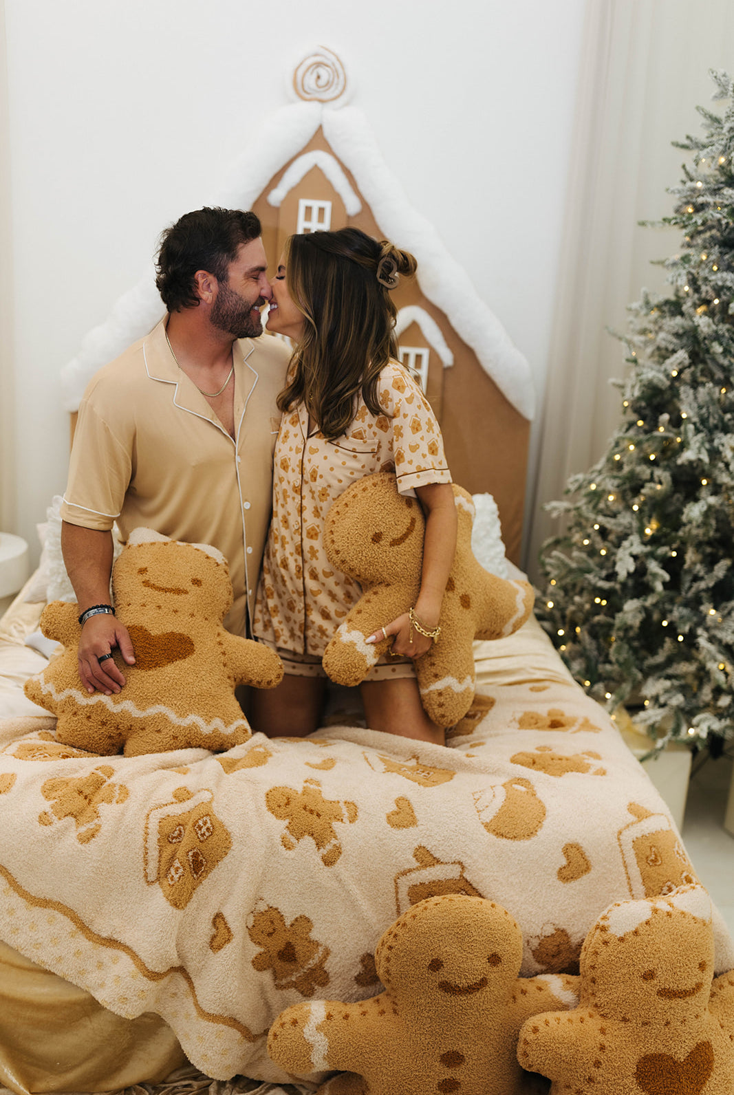 Couple sitting on a bed with gingerbread-themed decor, including a gingerbread house and tree.