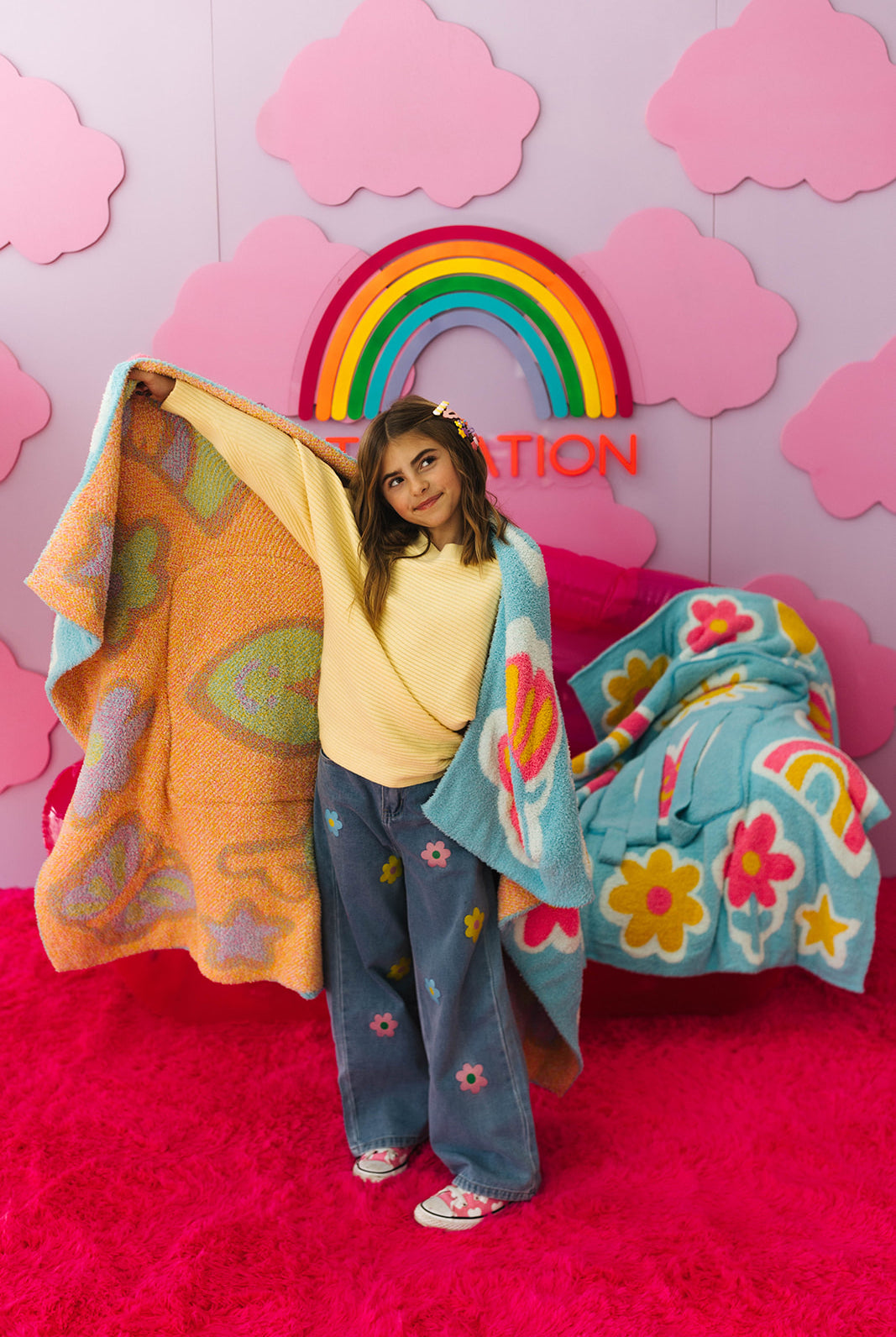 Person holding colorful blankets against a pink wall with a rainbow and clouds.