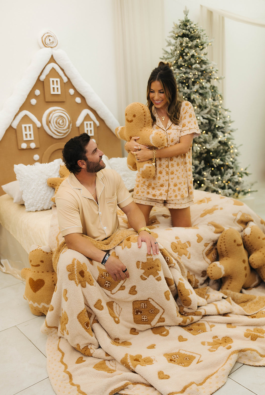Man and woman in pajamas sitting on a bed with gingerbread-themed decor and tree in the background.