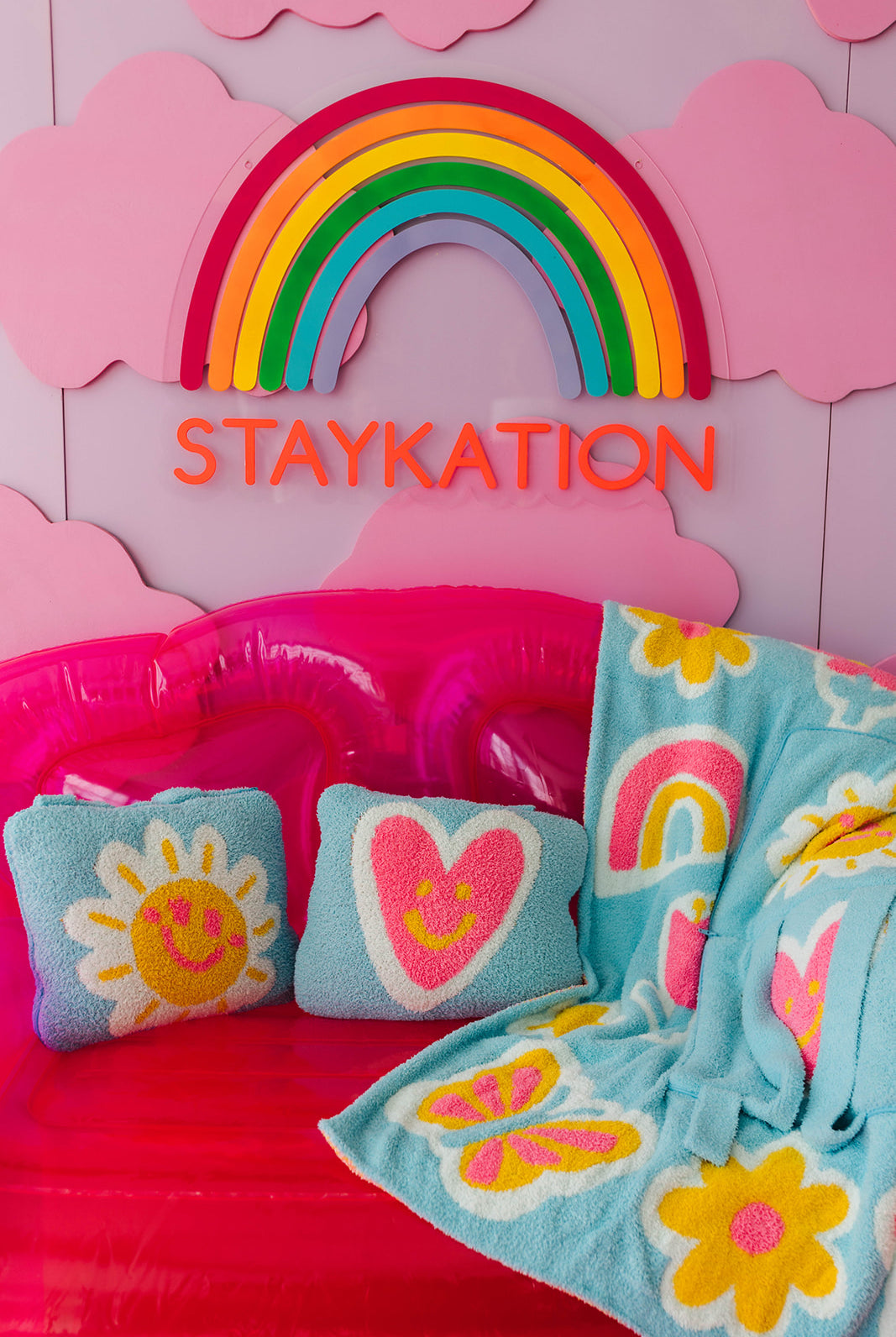 Children's inflatable bed with colorful pillows and blanket, featuring a rainbow and 'STAYKATION' text on a pink background.