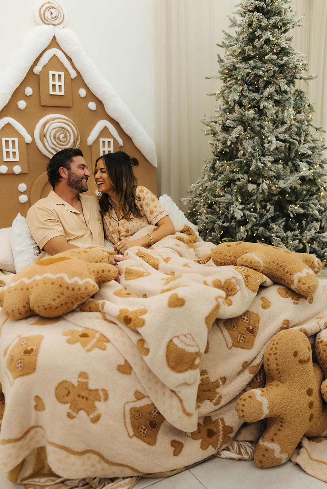 Couple under a gingerbread-themed blanket in front of a Christmas tree and gingerbread house.
