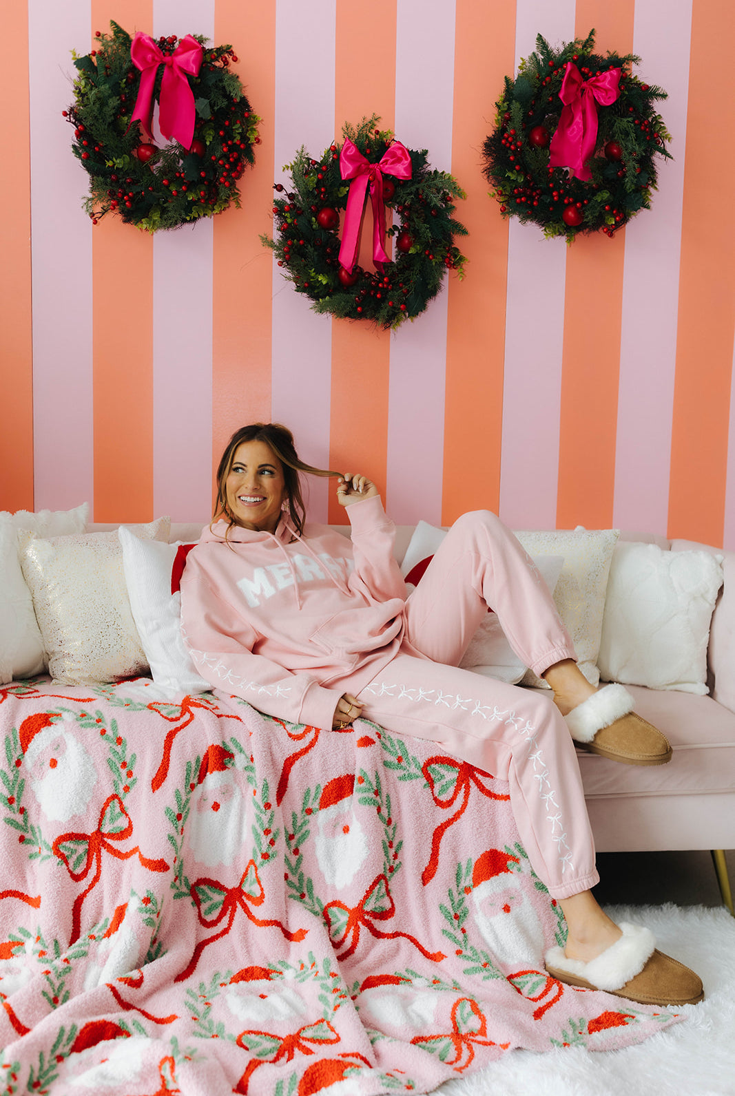 Woman in pink pajamas sitting on a couch with a Christmas-themed blanket and wreaths on a striped wall.