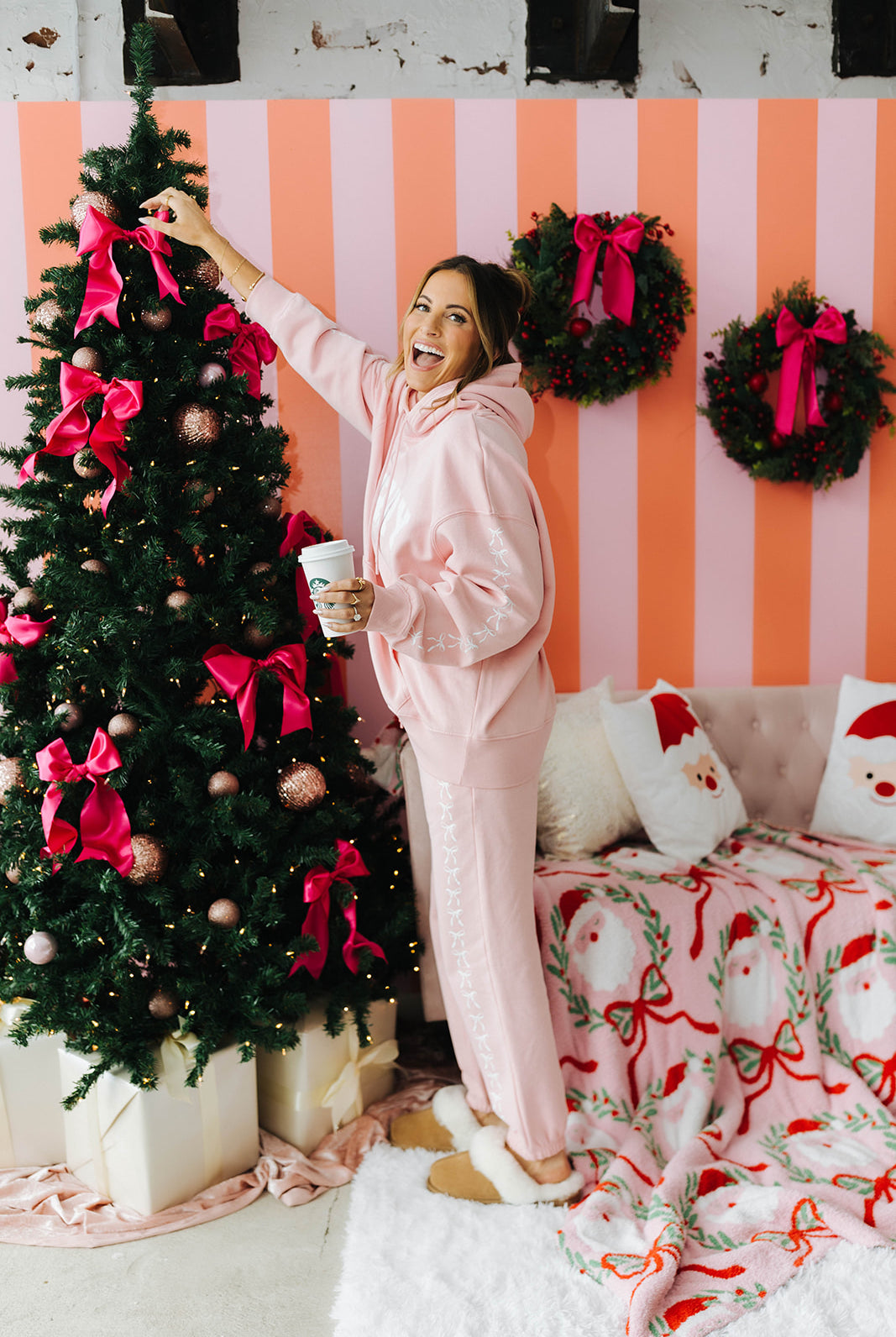 Woman in pink sweat set decorating a Christmas tree with a striped wall and festive decorations in the background.