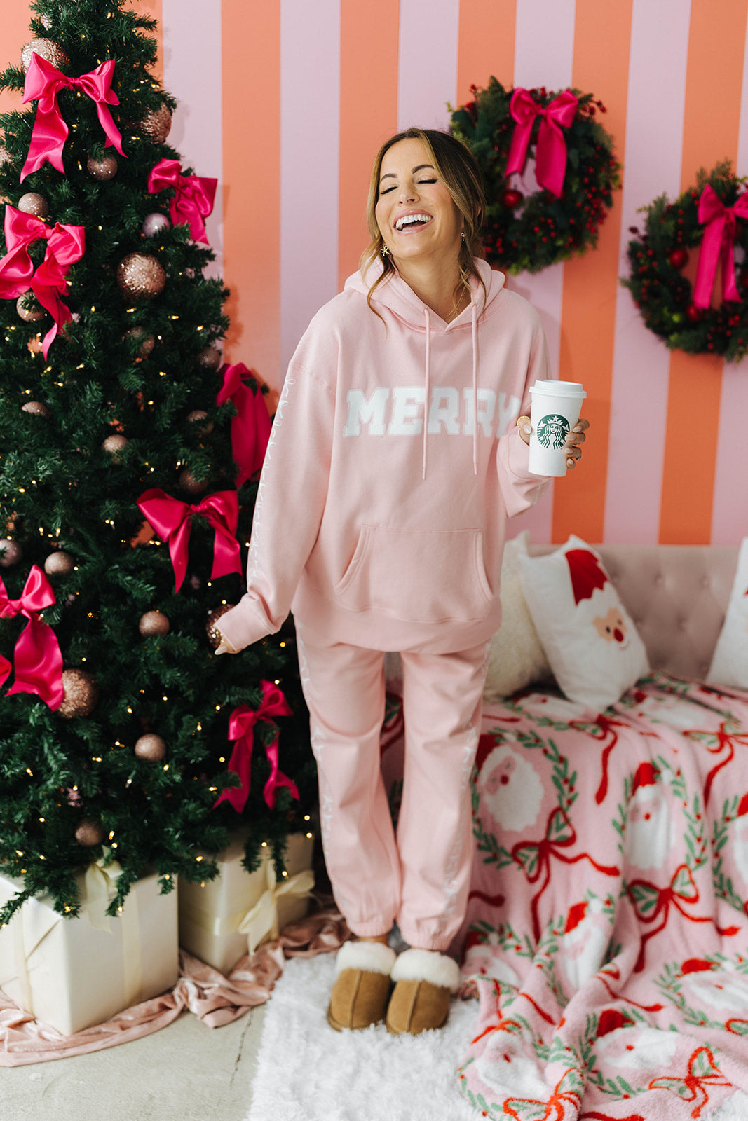 Person in a pink 'MERRY' hoodie holding a Starbucks cup, standing next to a decorated Christmas tree with pink bows and wreaths on a striped wall.