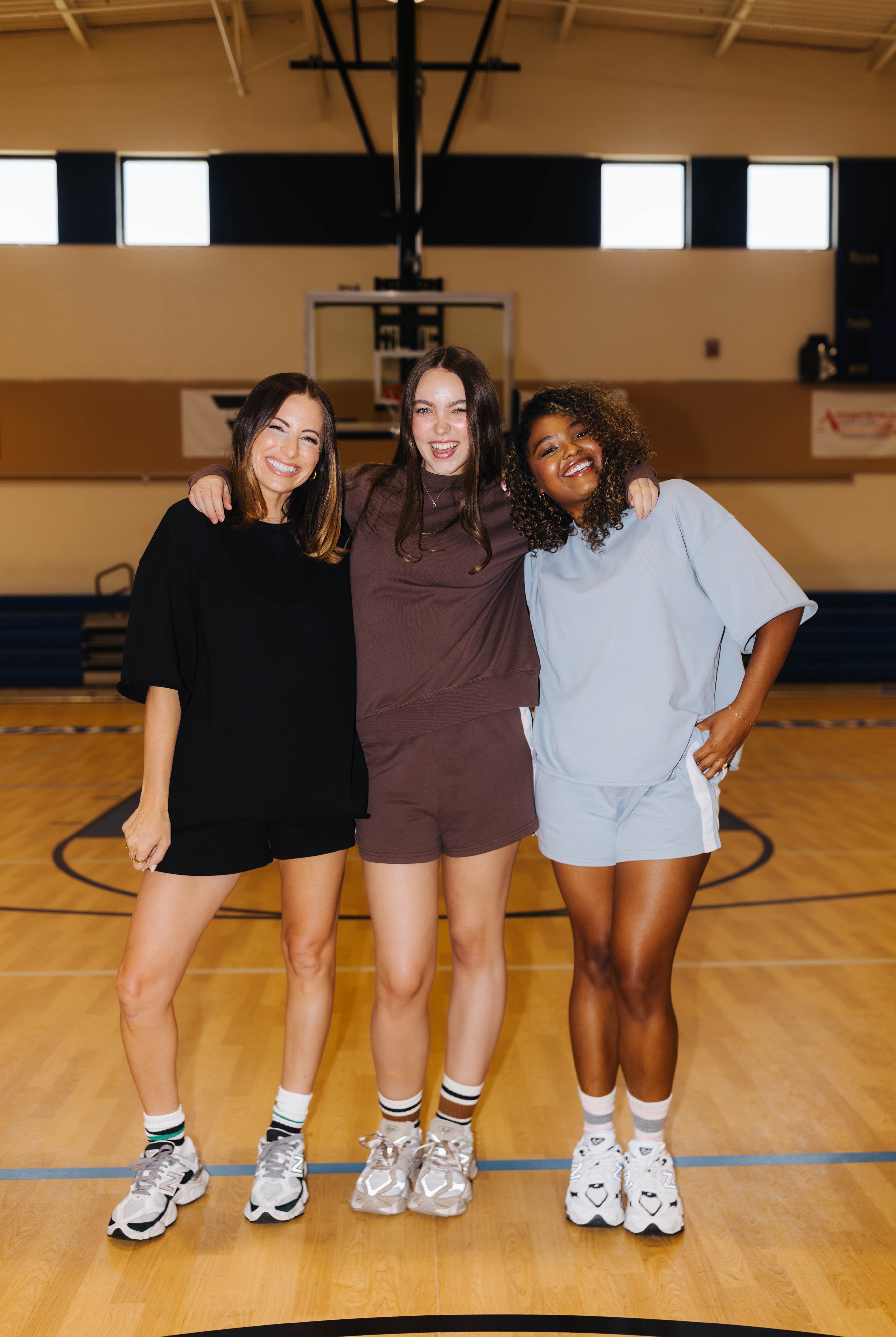 Three women posing together on a wooden basketball court, each wearing a matching oversized lounge set in black, brown, and light blue. The background shows a gymnasium with a basketball hoop and court markings.