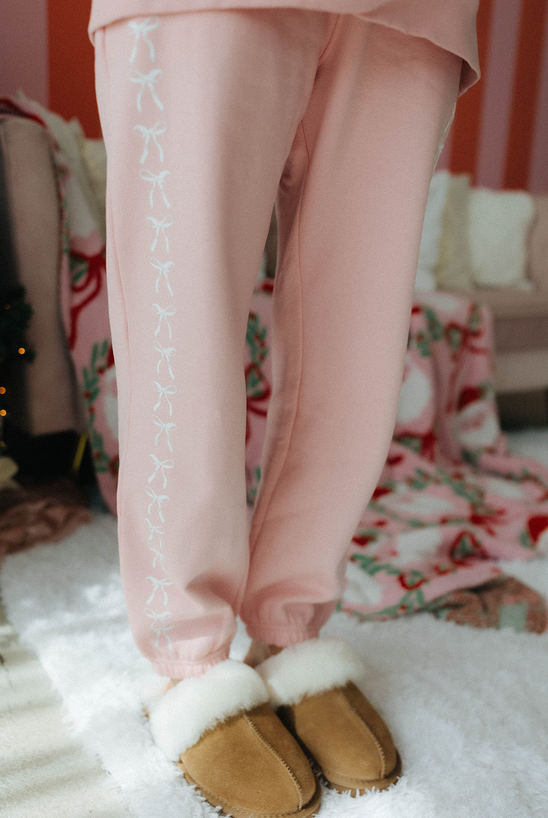 Person wearing pink sweatpants with white bows and brown slippers indoors.
