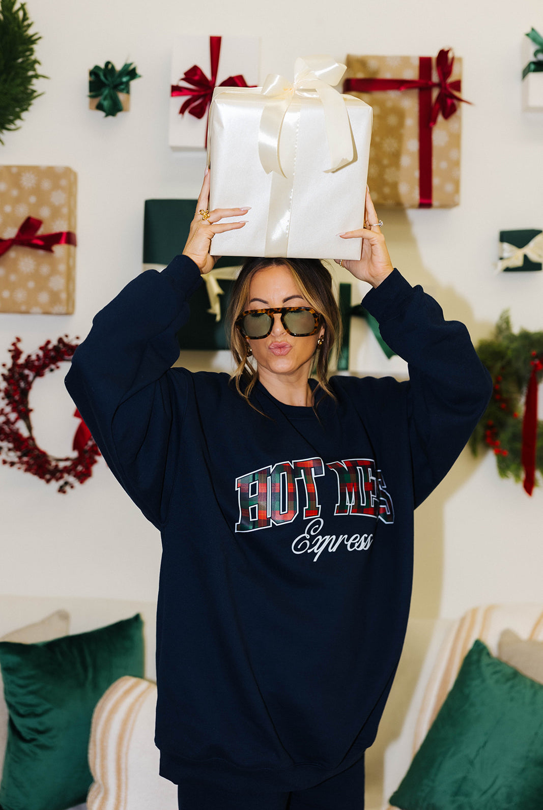 Person wearing a navy sweatshirt with 'HOT MESS Express' text, holding a gift box in a festive setting.