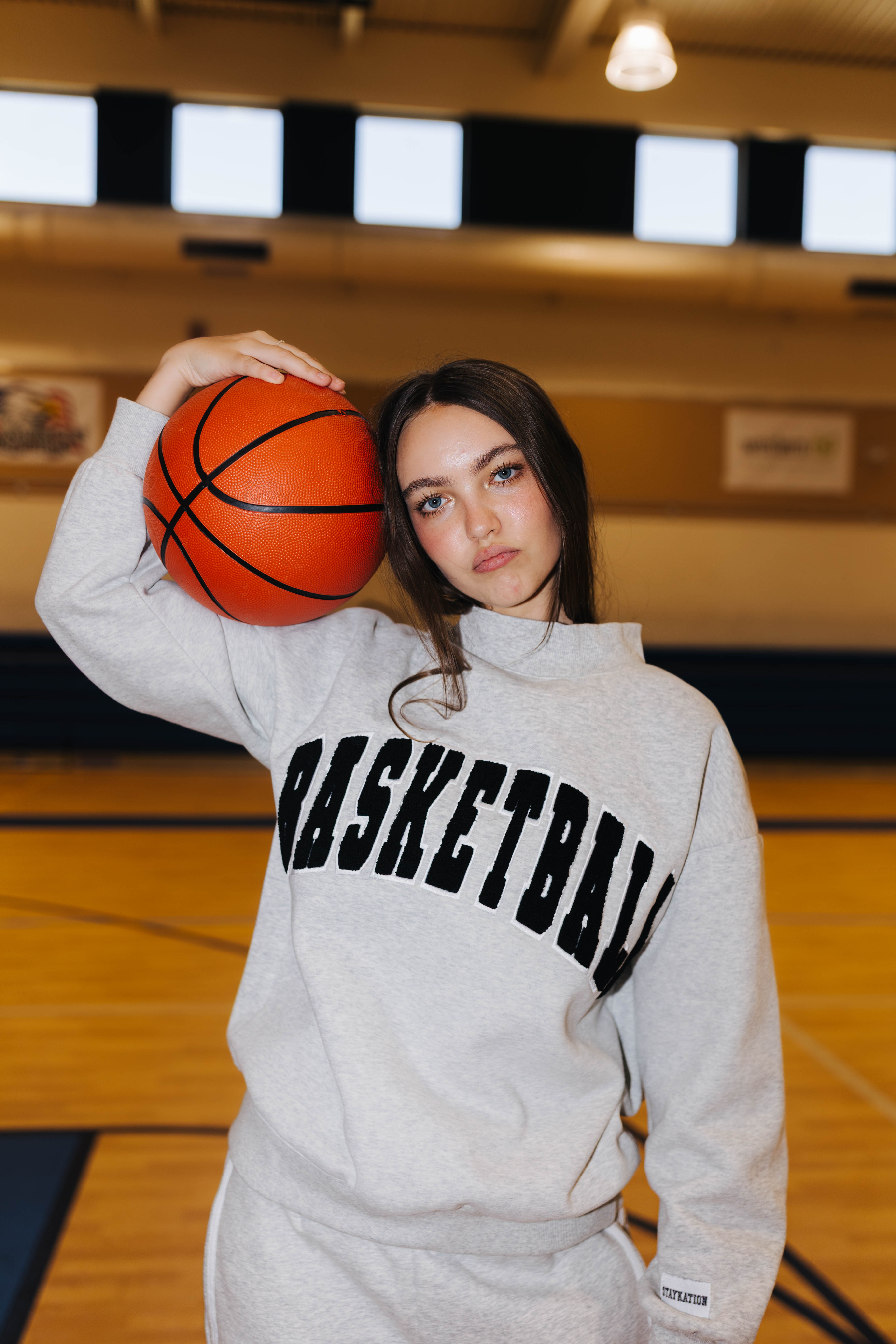 A young woman in a gym wearing a light gray sweatshirt with 'BASKETBALL' printed in black across the chest, holding an orange basketball against her shoulder.