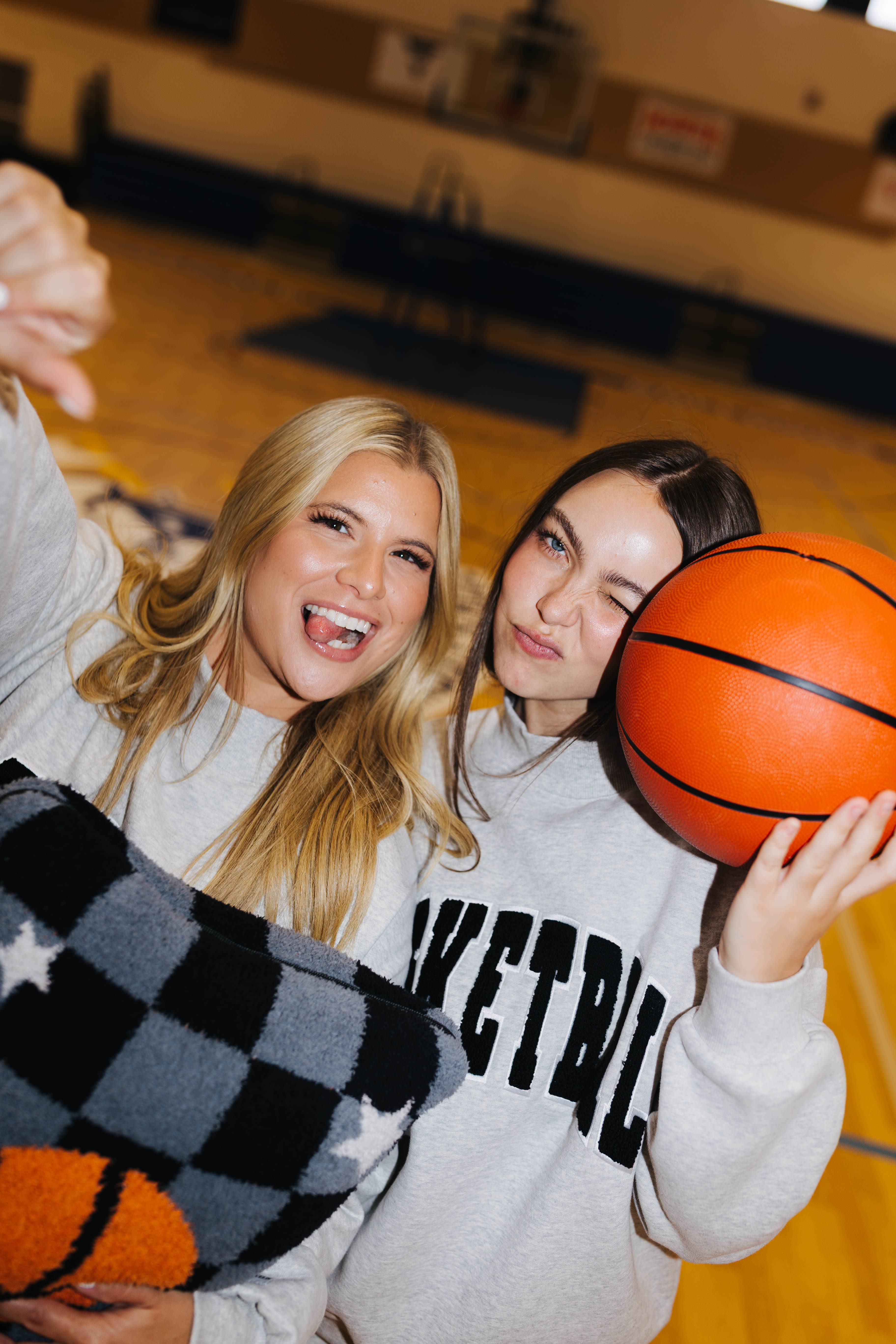Two women posing together on a basketball court, both wearing light gray sweatshirts with black lettering. One holds an orange basketball, and the other holds a black, gray, and white checkered quillow.
