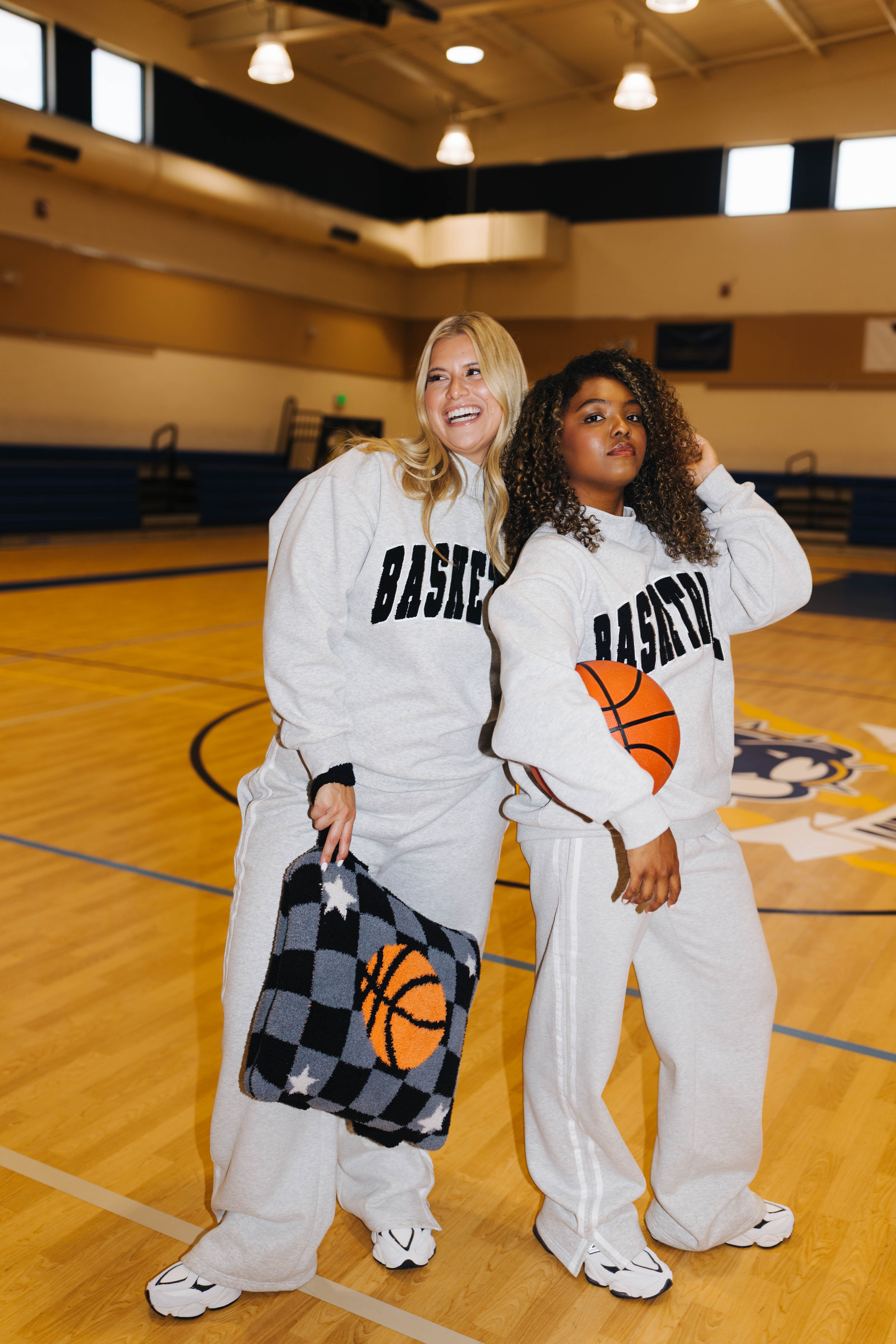 Two women posing on a basketball court wearing matching sweatpants and sweatshirts with 'BASKETBALL' printed in black.
